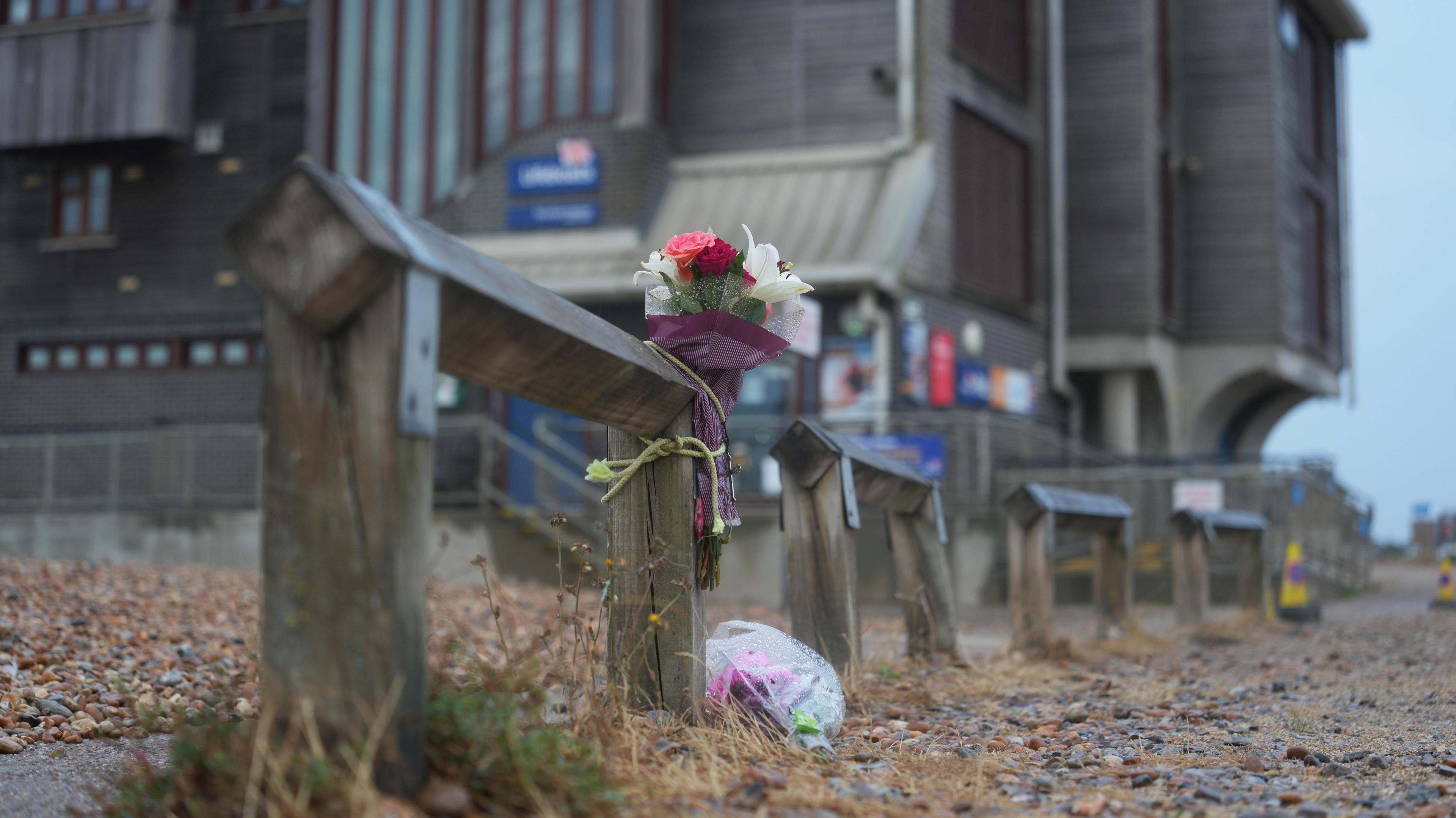 A bouquet of flowers is tied to a wooden beach barrier on a pebble shoreline near Shoreham RNLI. Buildings and seafront structures can be seen in the background.