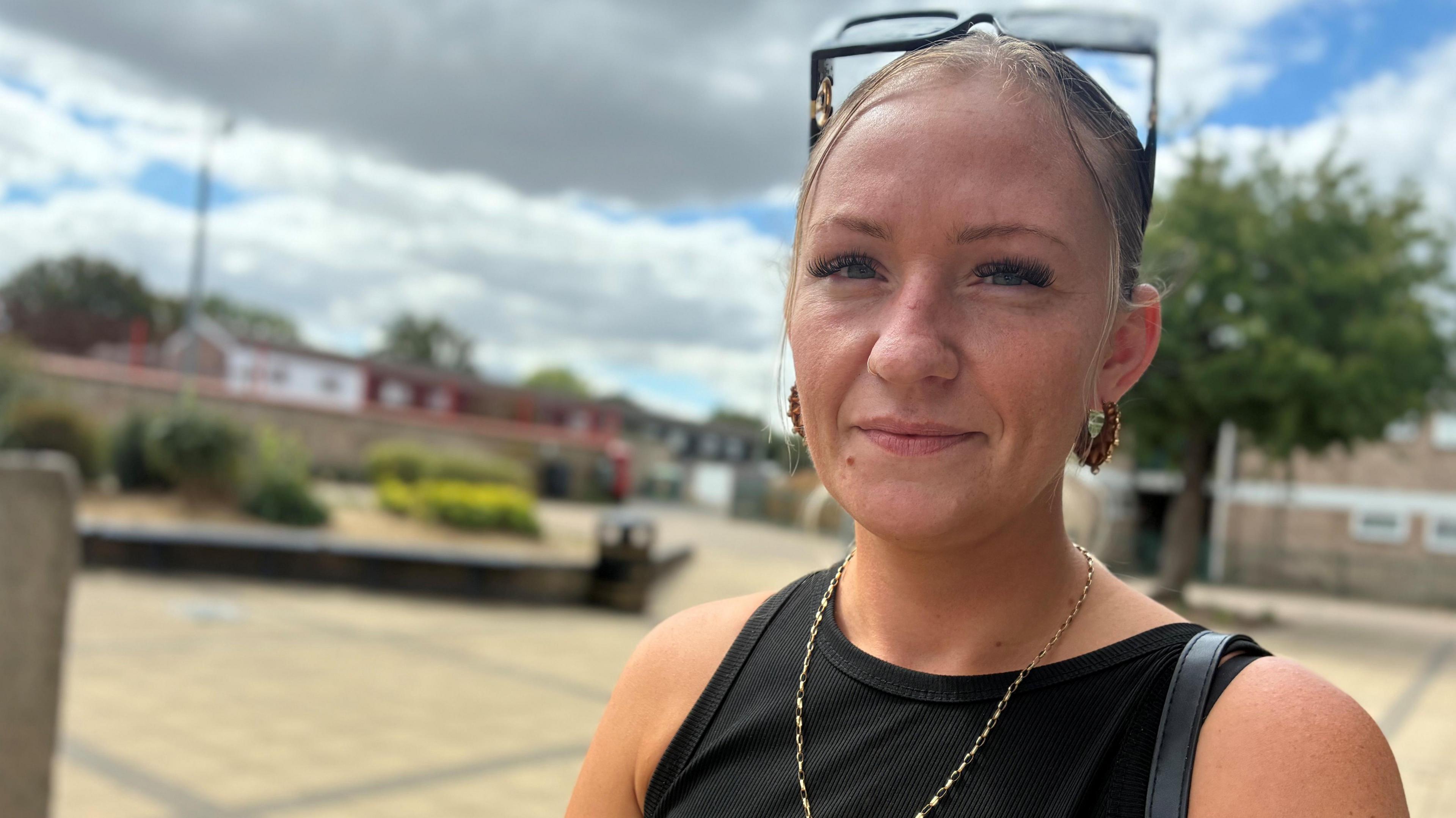 A woman with sunglasses on her head is smiling at the camera. Behind her is a housing estate.
