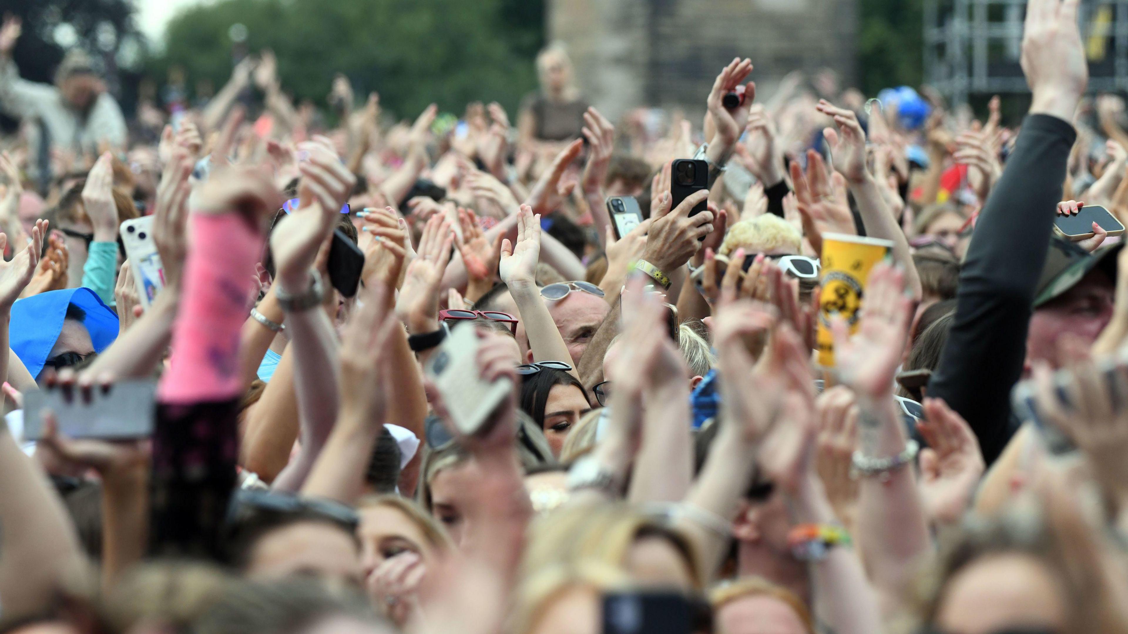 Festival-goers at event. People in the crowd are waving their hands in the air. Lots of people are holding their phones while others are holding up cups.