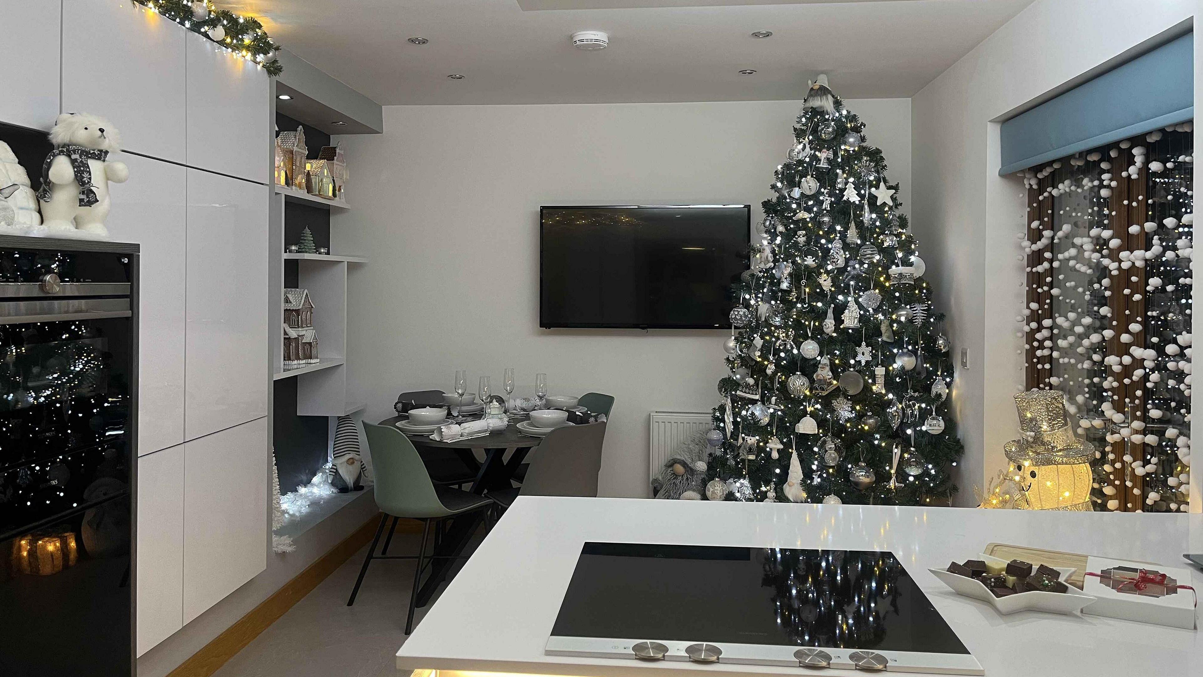 A large Christmas tree sits in the corner of a white kitchen. The tree is decorated with white Christmas baubles and white fairy lights. A light up snowman in a top hat stands in front of patio doors, which are decorated with a cascading wall of baubles.  A polar bear wearing a scarf perches on  top of the oven. 