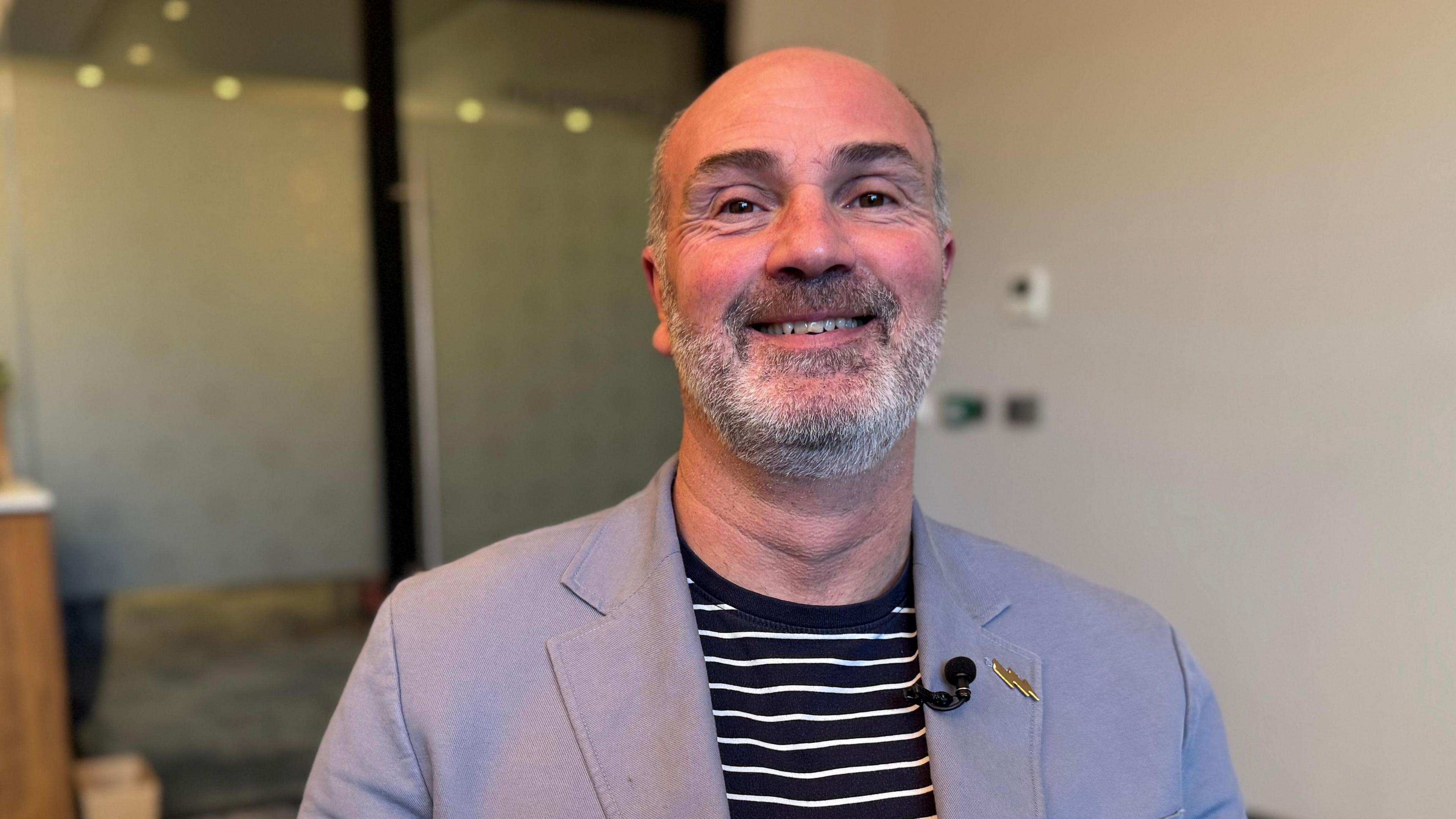 Mae Llŷr Morus yn gwisgo siaced llwyd gyda chrys streipiog. Mae ganddo farf.