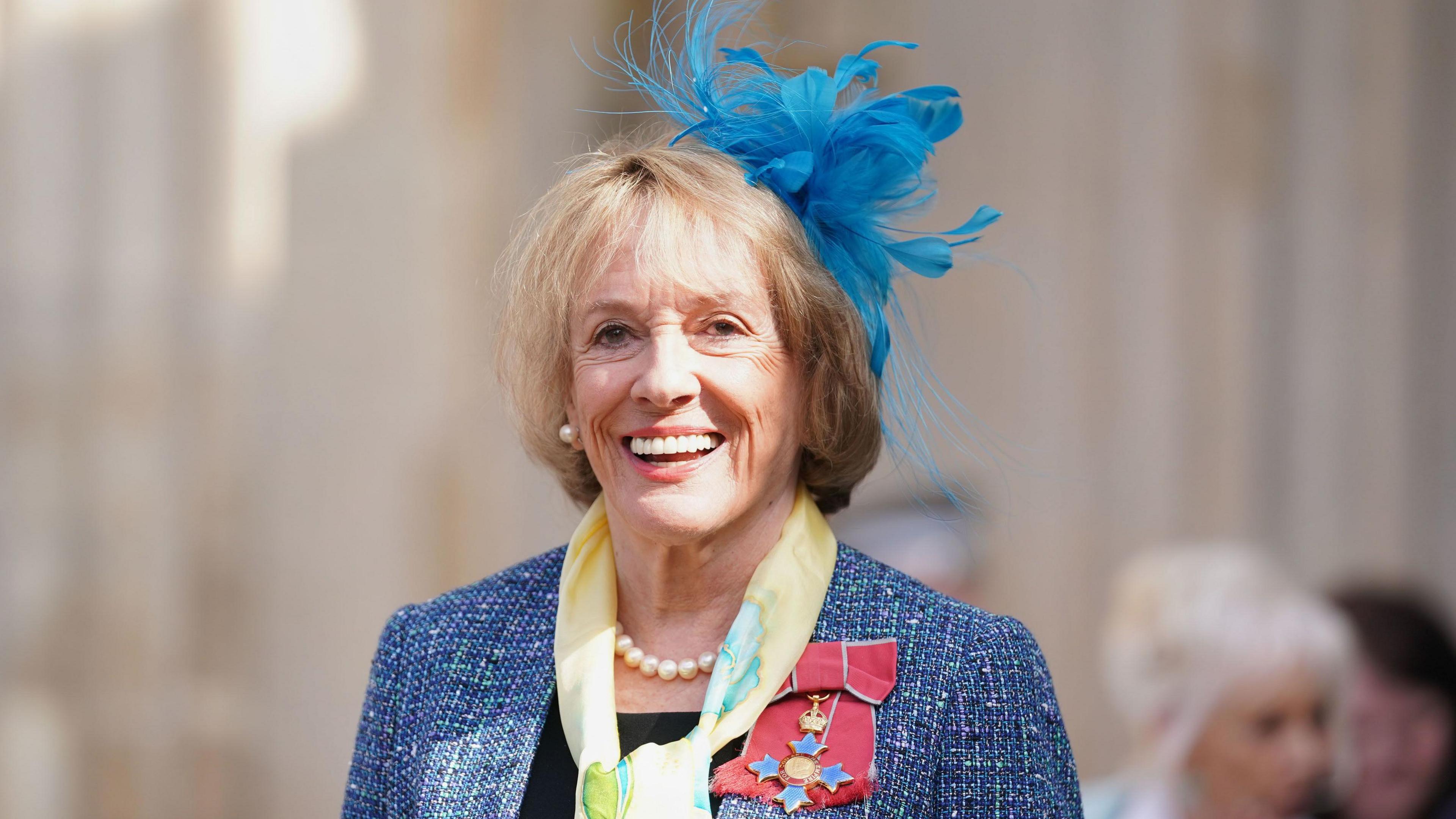 Dame Esther Rantzen, wearing blue headwear, a pearl necklace, scarf and badges, with a blue blazer and dark shirt, smiling towards a camera with the sun on her face. 