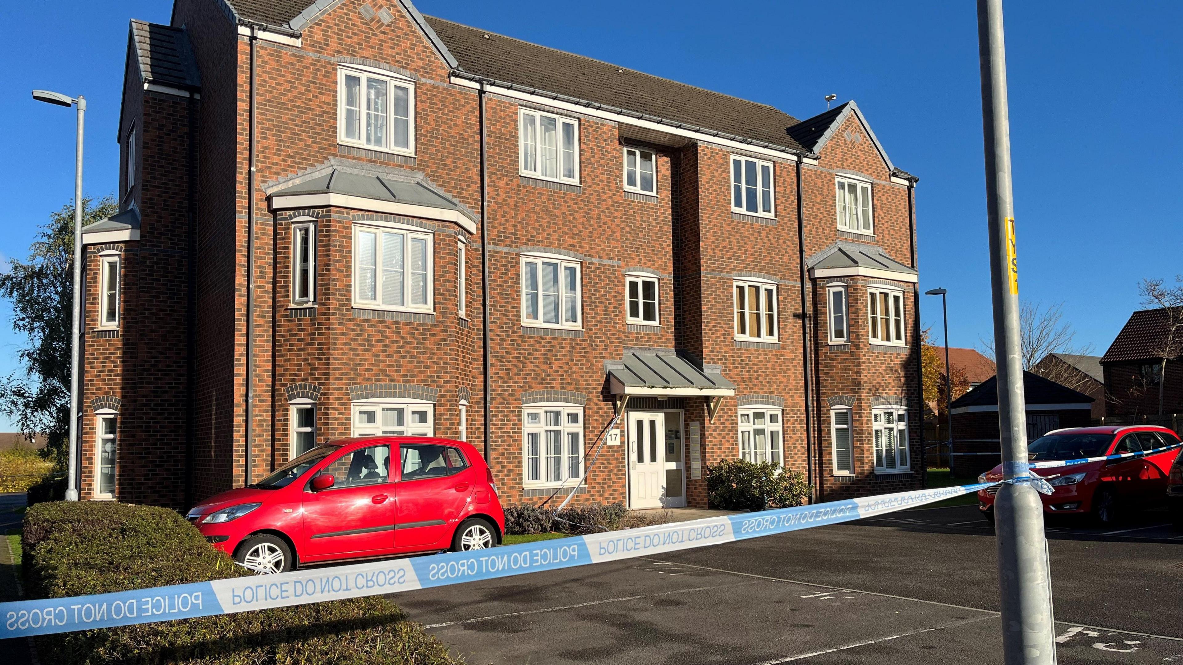 A  three-storey red brick flat building with police tape attached to a lamppost outside. There is a carpark in front of the building with two red cars parked. The sky is blue and clear.