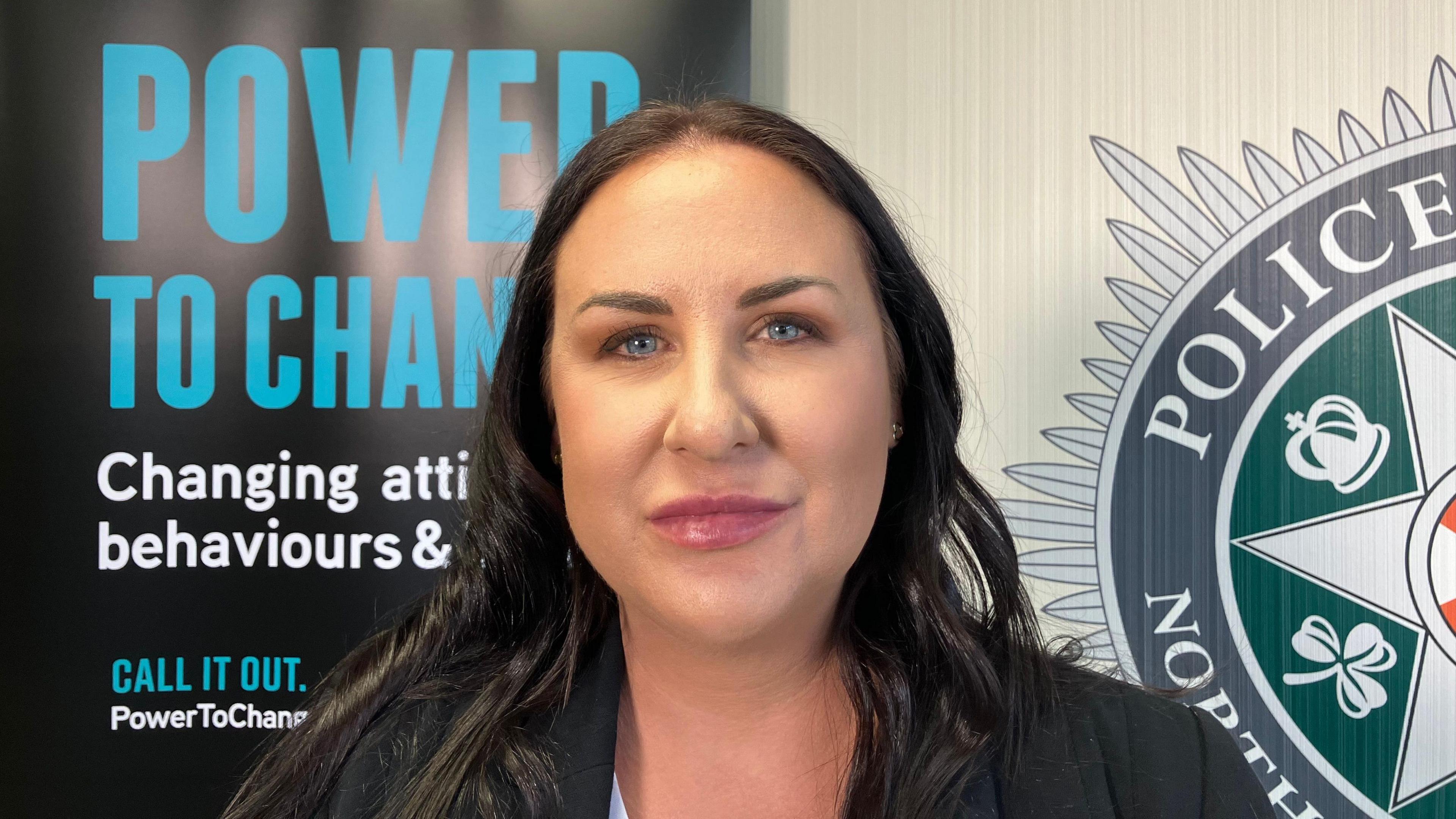 A woman with long brown hair, wearing a black blazer. She is looking at the camera. Half of a PSNI logo is visible on the right hand side of the wall behind her. To the left is a black poster reading "Power to change" in blue. 
