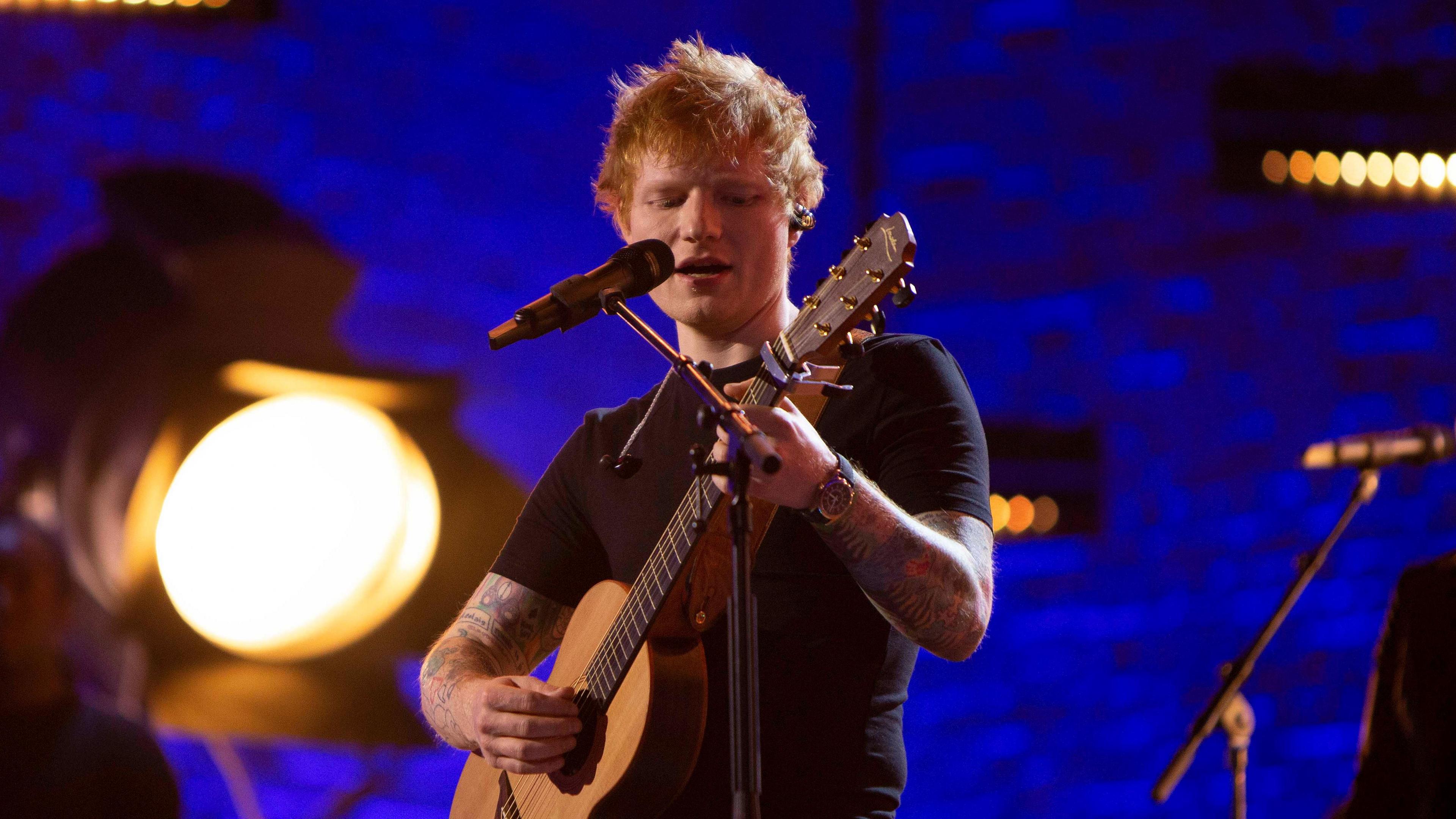 Ed Sheeran singing at a microphone and playing an acoustic guitar. A Large stage light in the background.