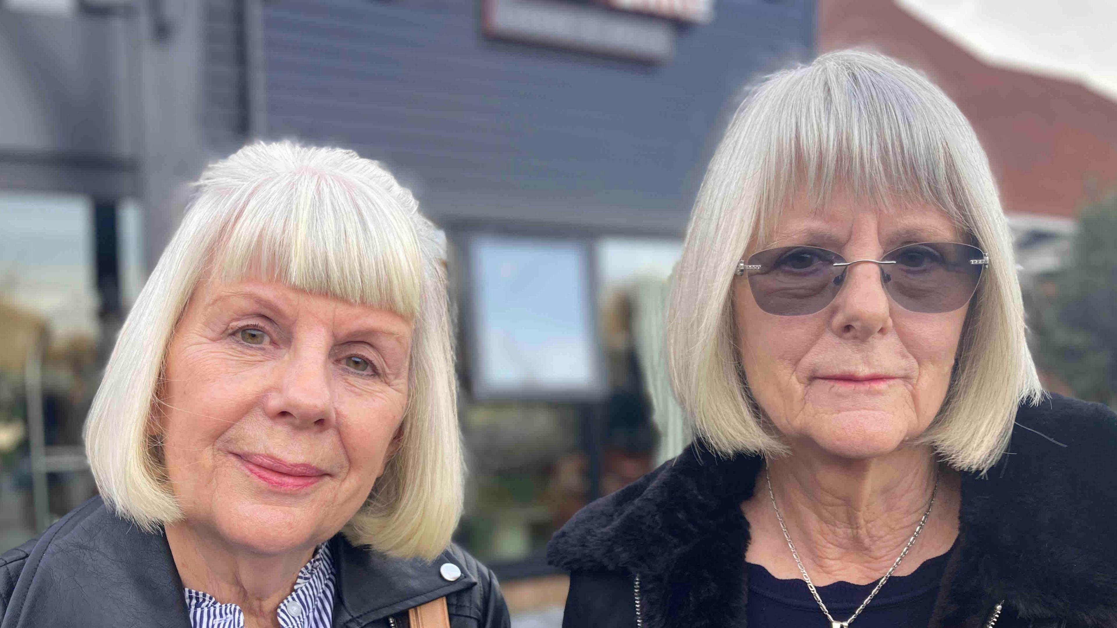 Penny Brind and Nicky Bettridge both have light-grey hair in a bob style with a fringe. Ms Brind wears a leather jacket over a striped top and Ms Bettridge, who is taller, has dark glasses and a dark blue jacket or coat.