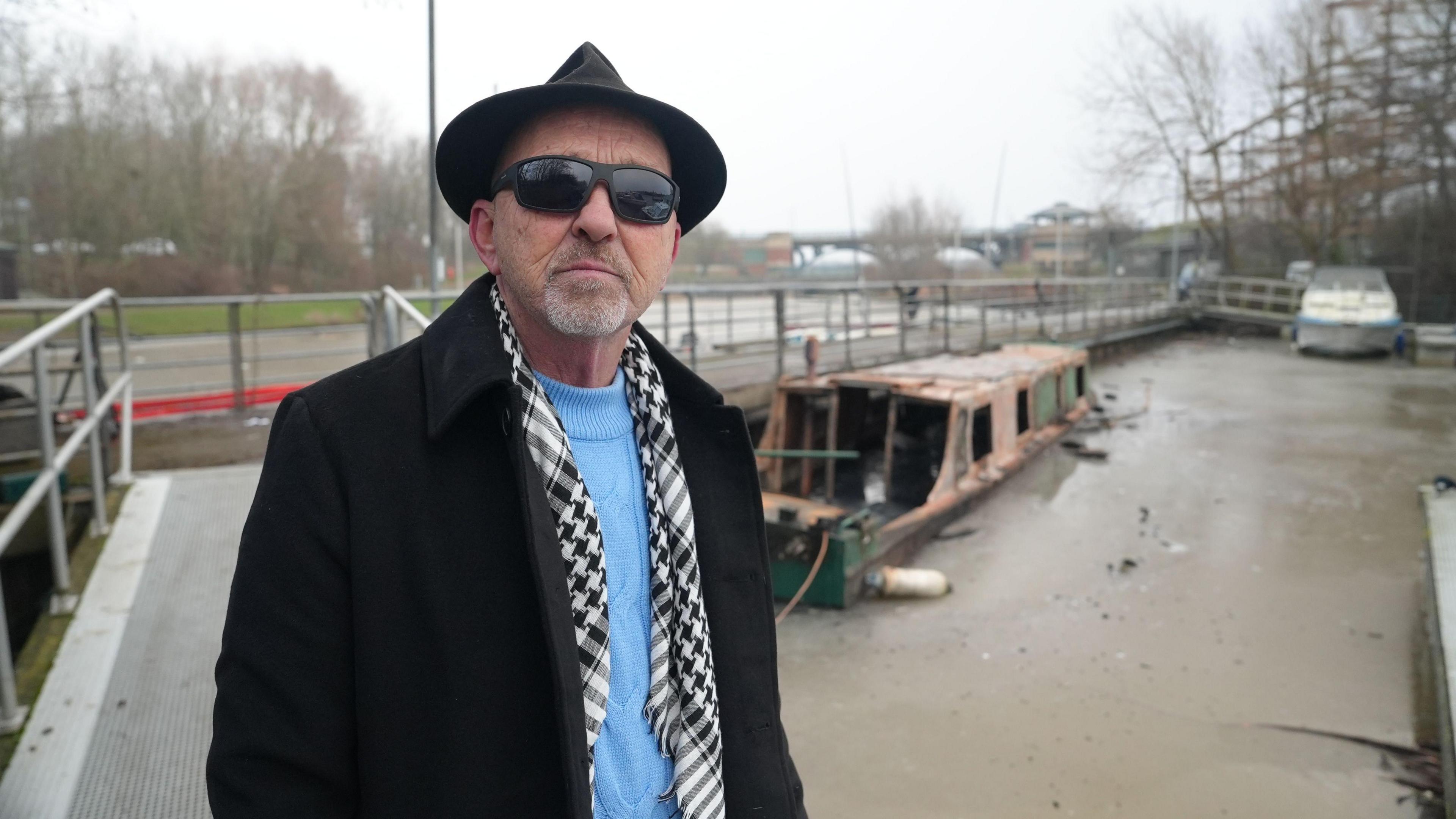 Graham Bennett looking into the camera  in front of his fire damaged barge. He's wearing black sunglasses, a black hat, black and white scarf, light blue jumper and black coat.