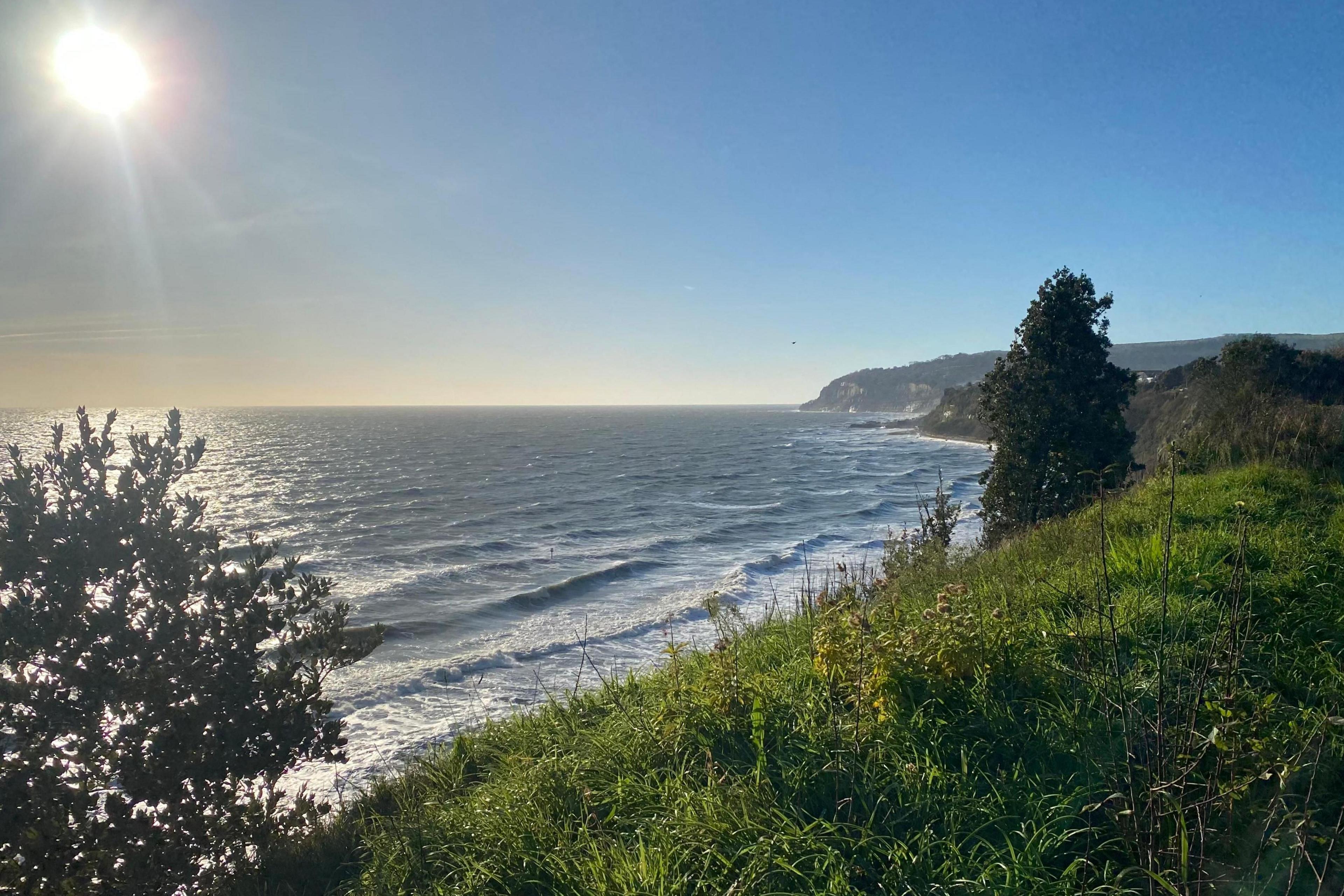 A sunny coastline shot with cliffs looking down over waves coming in. A clear blue sky has the sun sat in the top corner of the image.