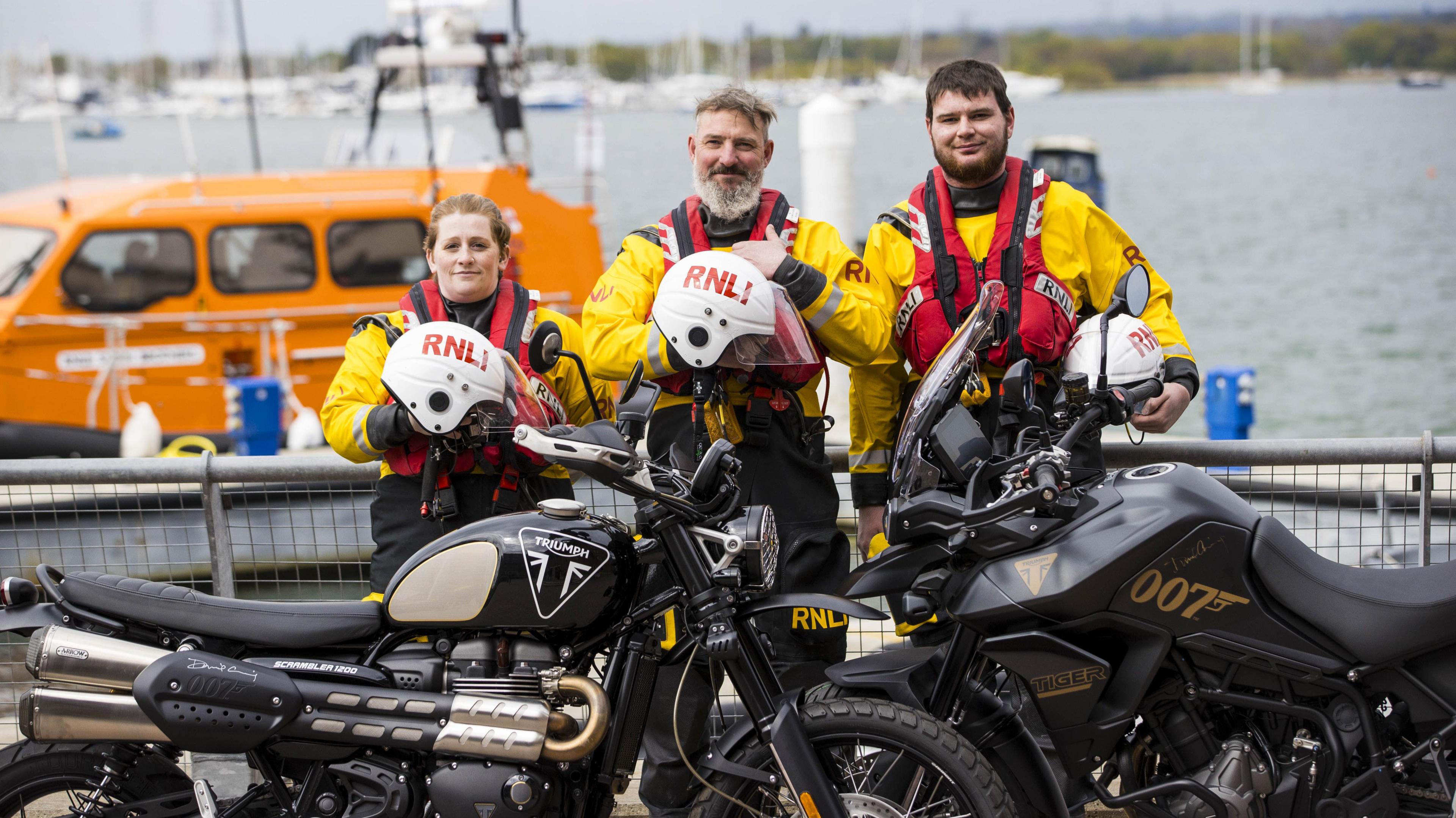 Three members of a lifeboat crew, a woman and two men, stand behind two motorbikes. There is a lake and boats behind them.