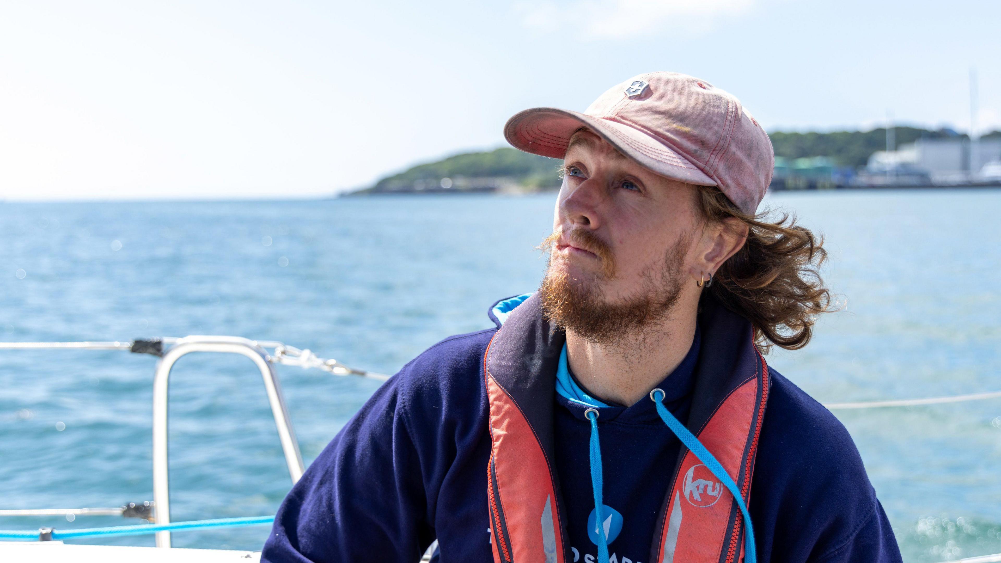 A man with long flowing hair, a beard and a pink cap sits on a boat, looking away from the camera. He is wearing a life jacket and a blue hoodie.