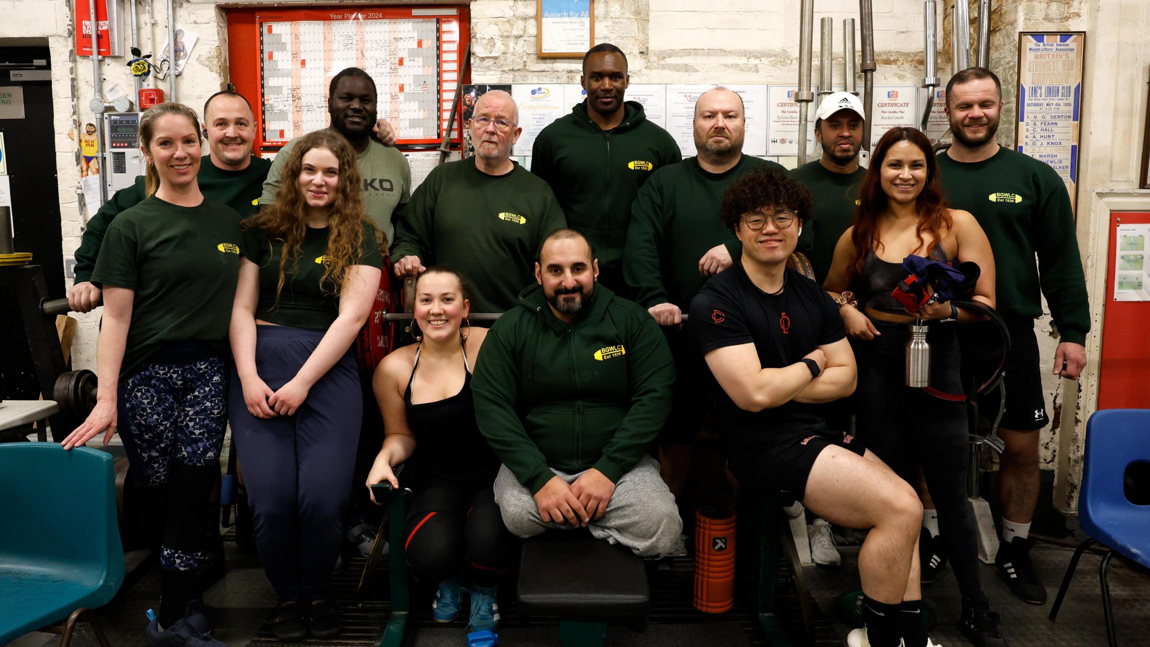 A group of 13 men and women are gathered for a group picture. Some are sitting in the front while others stand at the back within the gym.