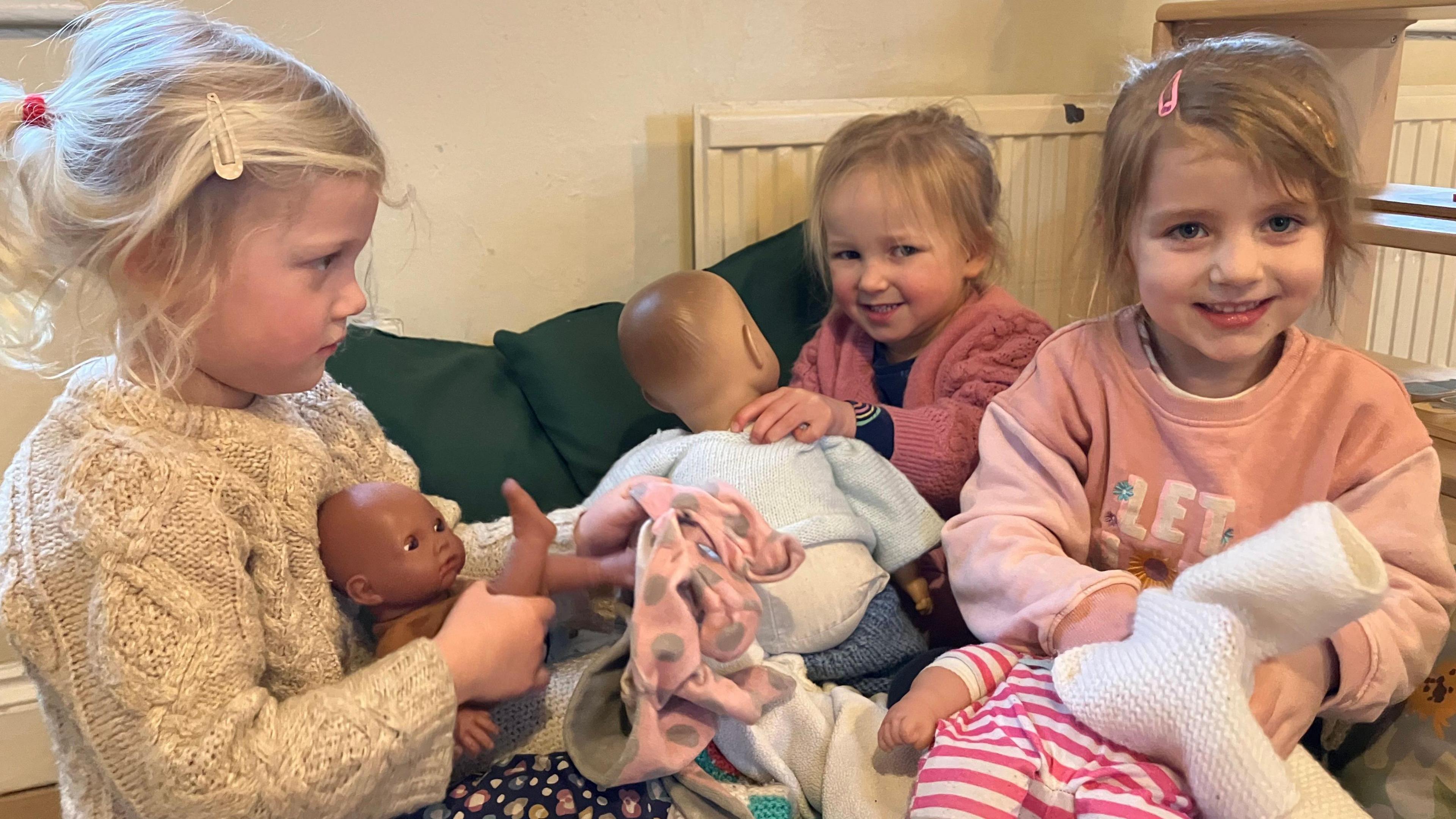 Three little girls sit in a circle playing with dolls and dressing them. 