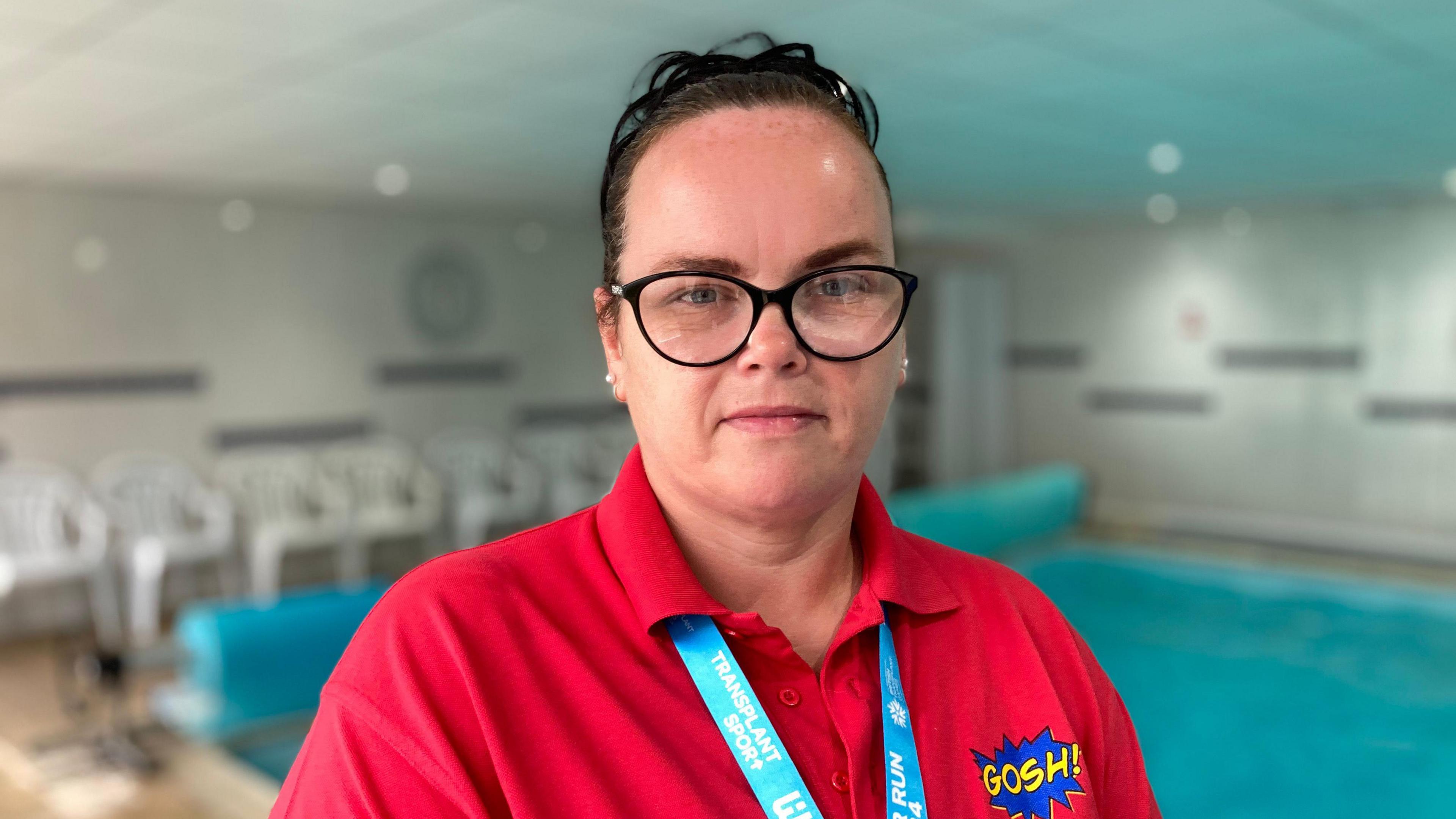 Sandra Tomlinson in a red t-shirt and glasses, looking into the camera and standing next to a swimming pool.