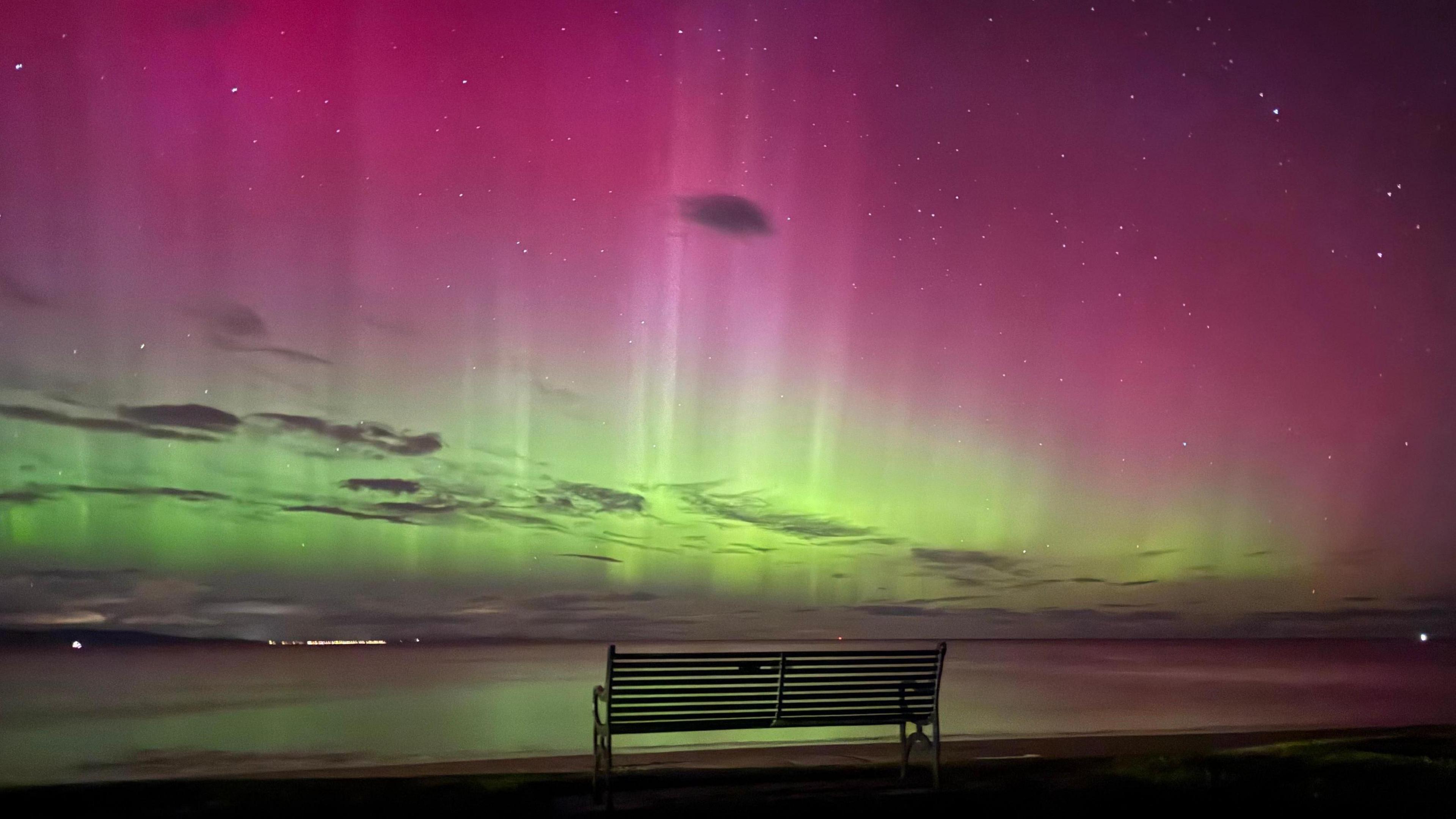 A bench sits in front of the sea in the dark. It is illuminated by a bright green and pink sky, which is dotted with dark clouds. The water reflects from the green lights
