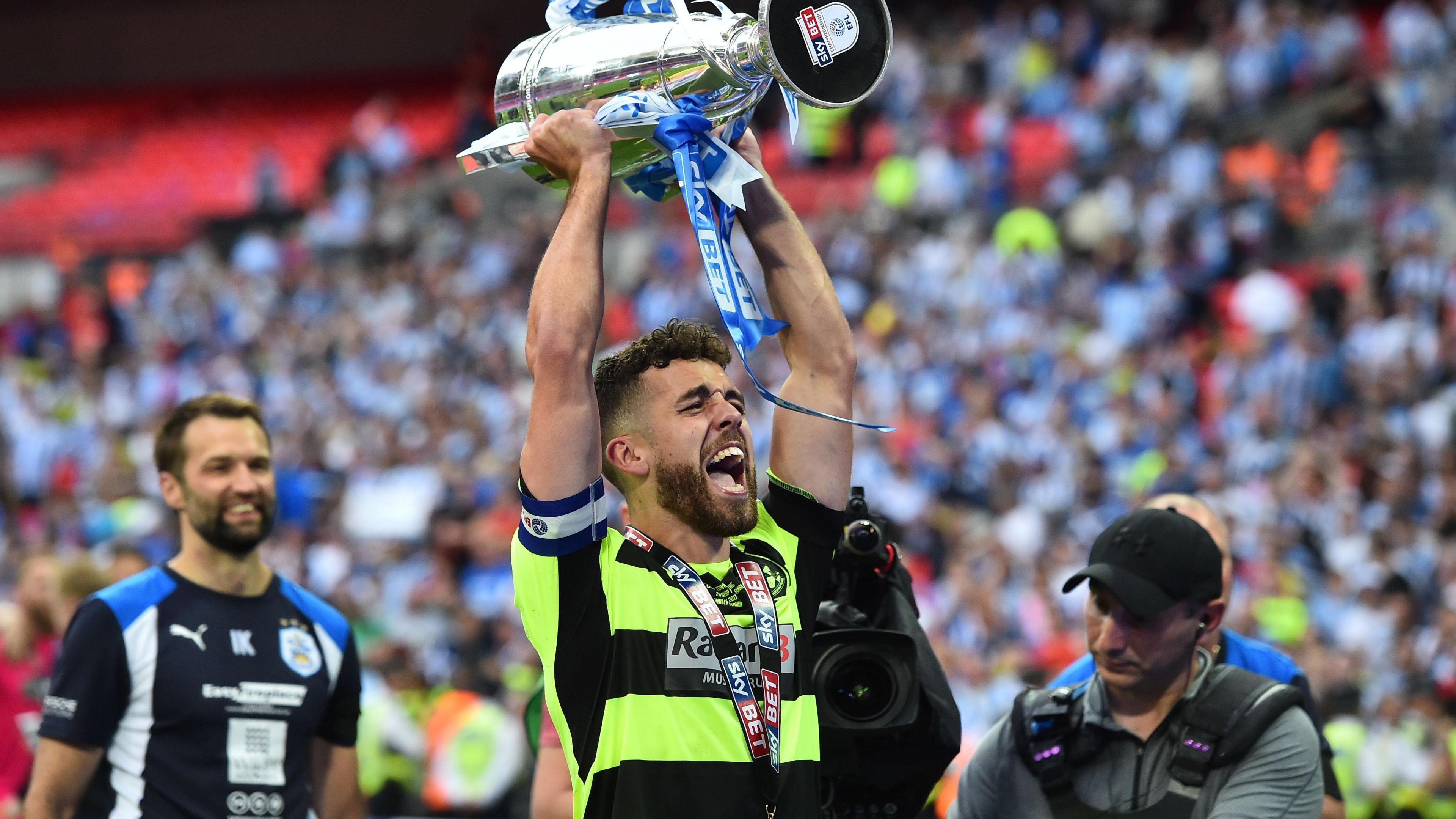 Tommy Smith, wearing a fluorescent green and black hooped Huddersfield away shirt, lifts the Championship play-off final trophy at Wembley in May 2017, Blue and white shirts of the Huddersfield fans in the stands can be seen behind him.