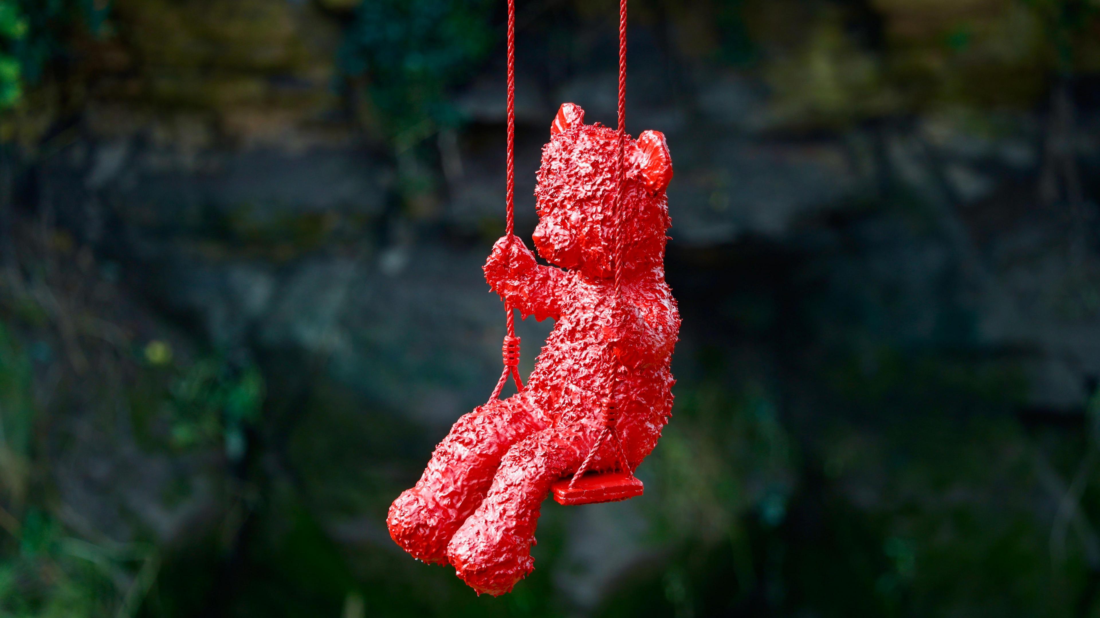 A sculpture of a bright red teddy bear on a red swing can be seen hanging from the bridge, pictured in close up against the blurry dark background of the riverbank. 