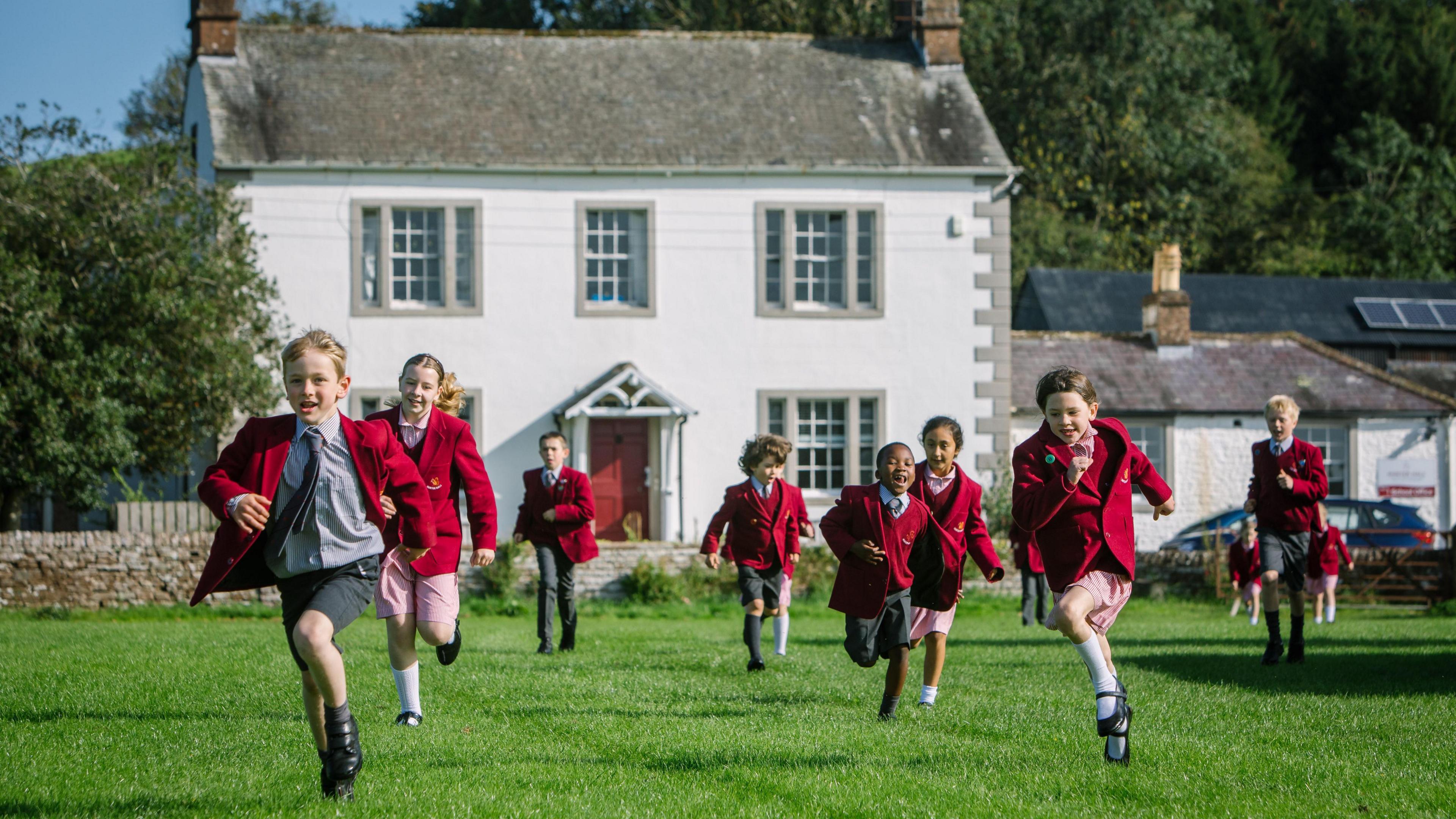 Almost a dozen boys and girls run across a grassed area in front of Hunter Hall School. They are wearing maroon blazers. The school building is an ornate two-storey structure painted white and has a grey roof.