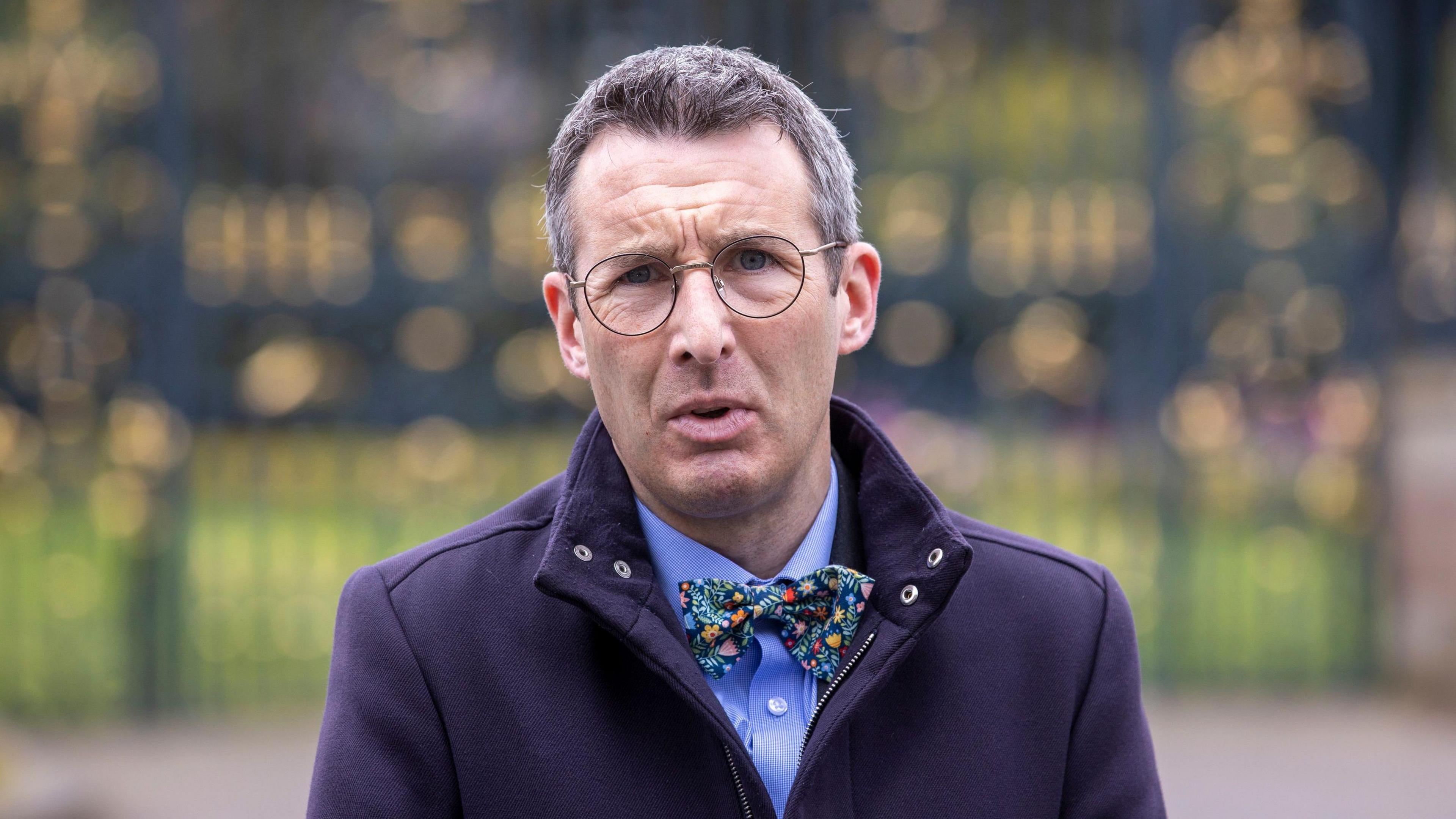 Andrew Muir standing with a long purple coat on with a bow tie and shirt. He is also wearing circular glasses standing outside.