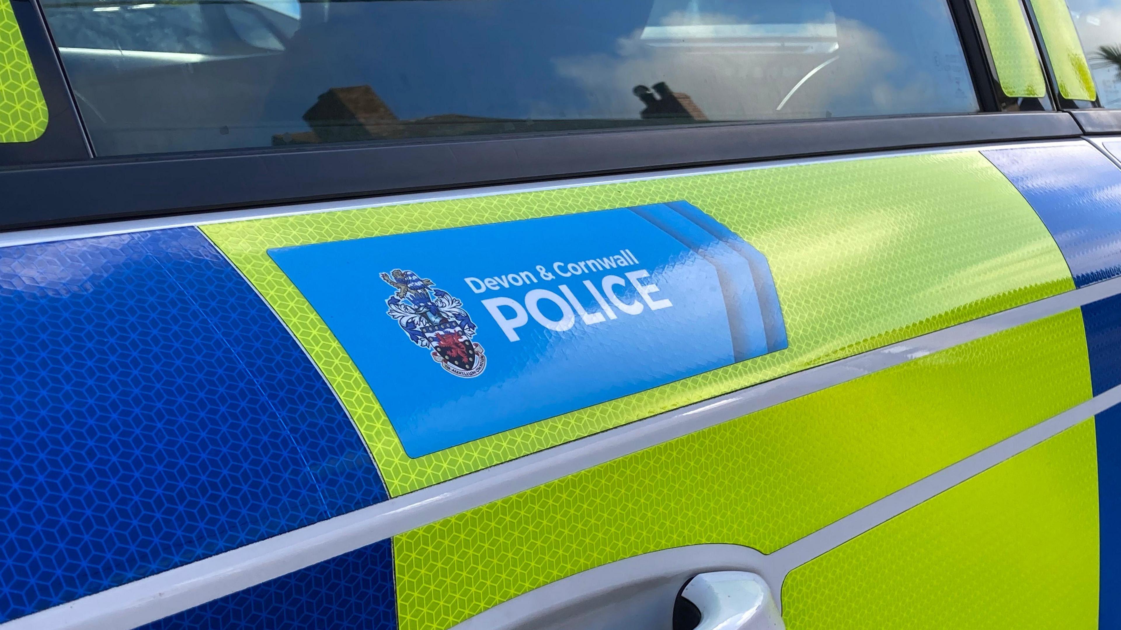 The side of a yellow and blue police vehicle. On the door, above the handle, has the a blue section with the words 'Devon and Cornwall Police'. There is a badge beside it.