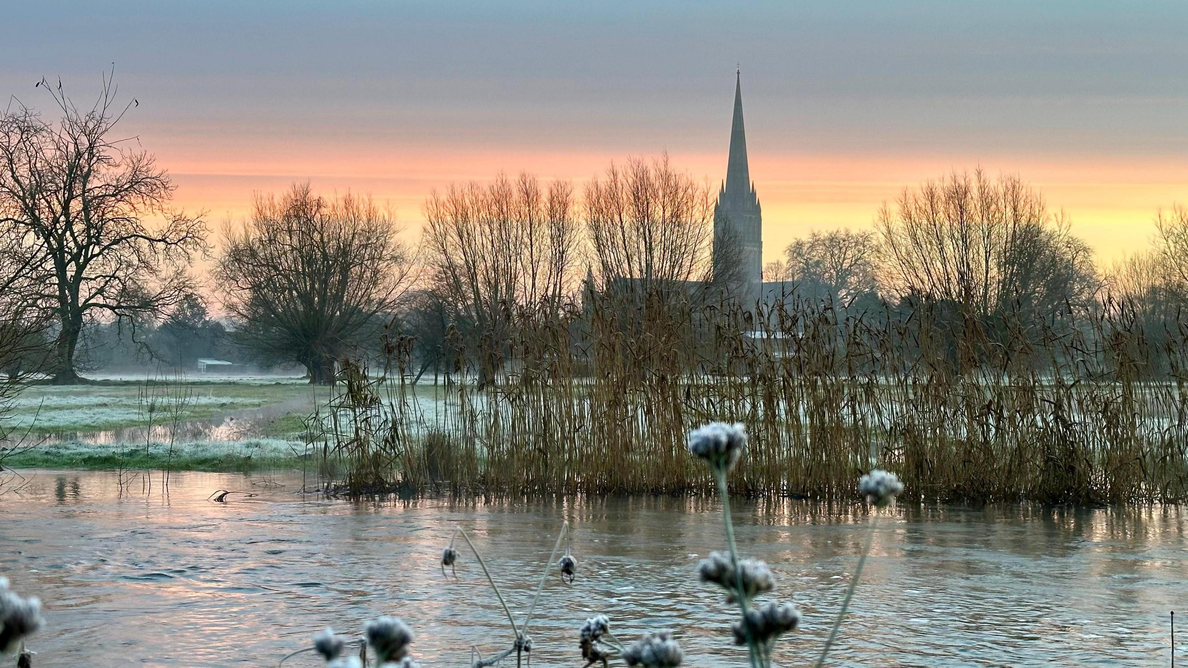 A winter scene in Wiltshire, Salisbury Cathedral in the background. There is a bit of a lake in front which is partially iced over. 