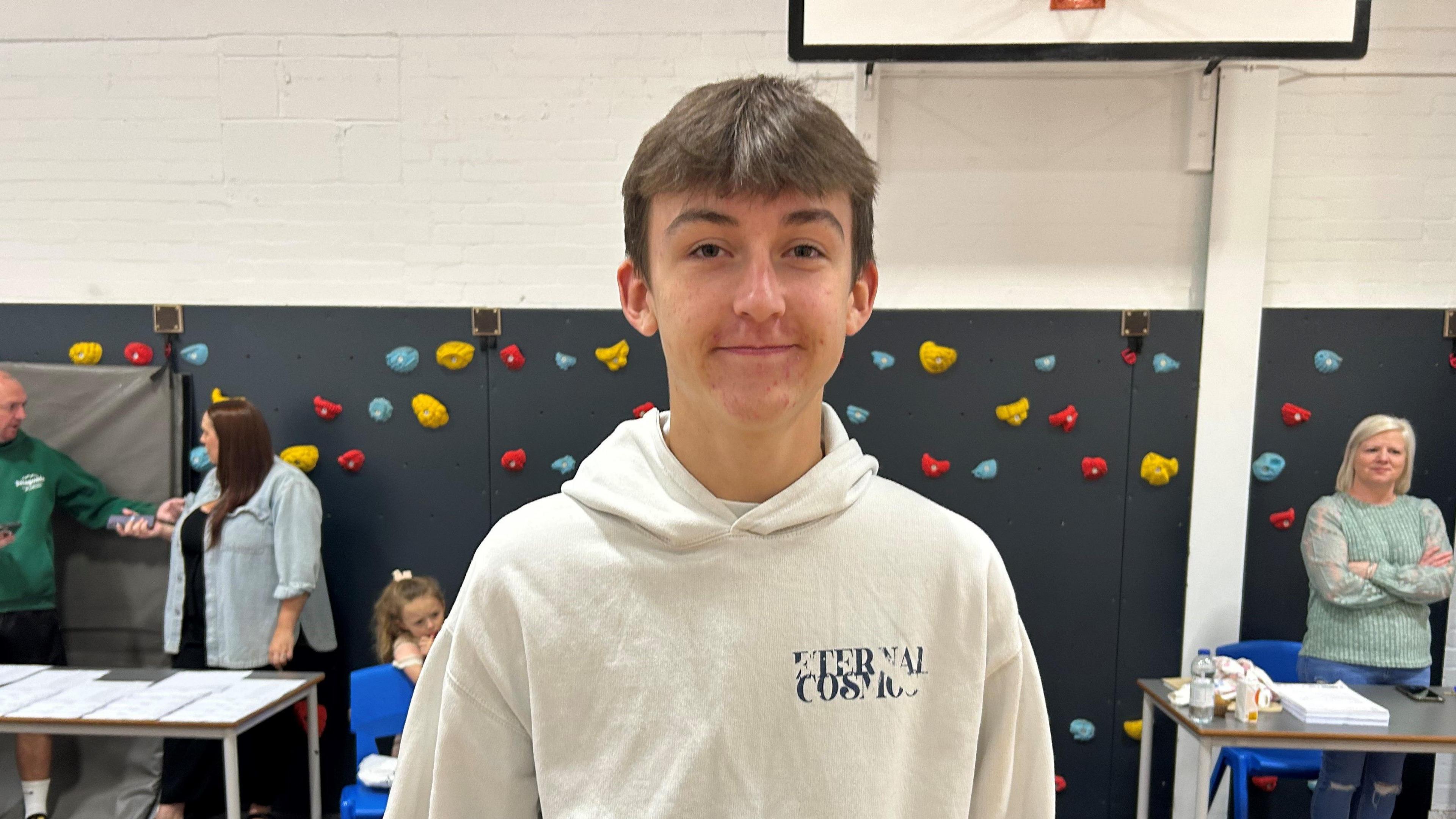 Sam stands in a sports hall holding his GCSE results. He is smiling, has short brown hair and is wearing a white hoodie. Behind him are people at tables with white envelopes laid out.