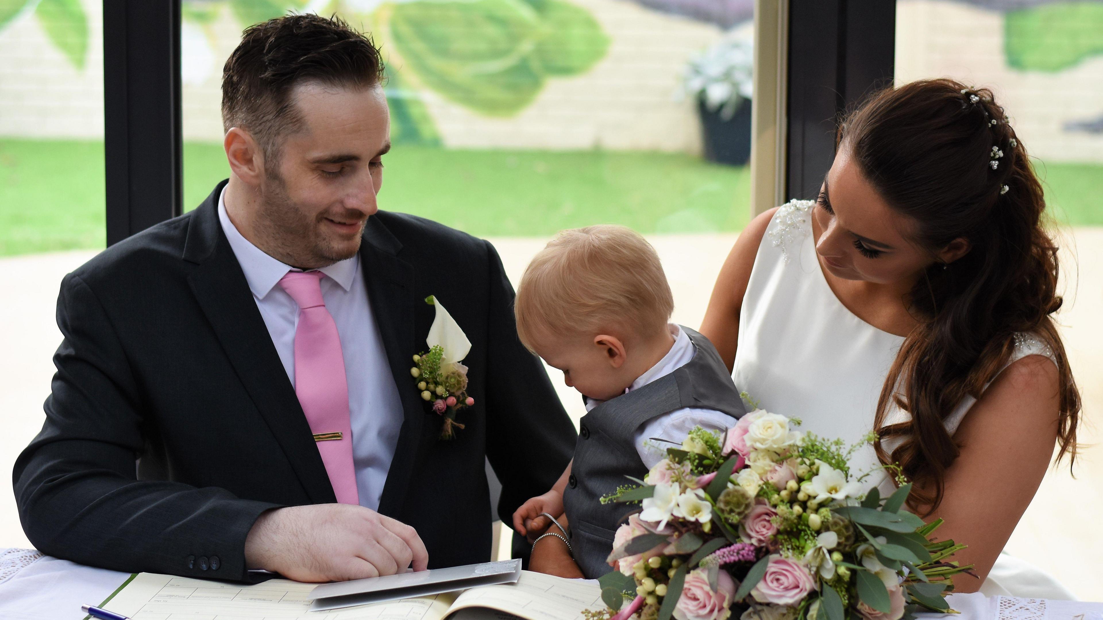 Gavin and Lori's wedding at the hospice. They're sat at a table with Fletcher between them, on Lori's knee. Gavin is wearing a suit and Lori's wearing a wedding dress. There's a bouquet on the table.