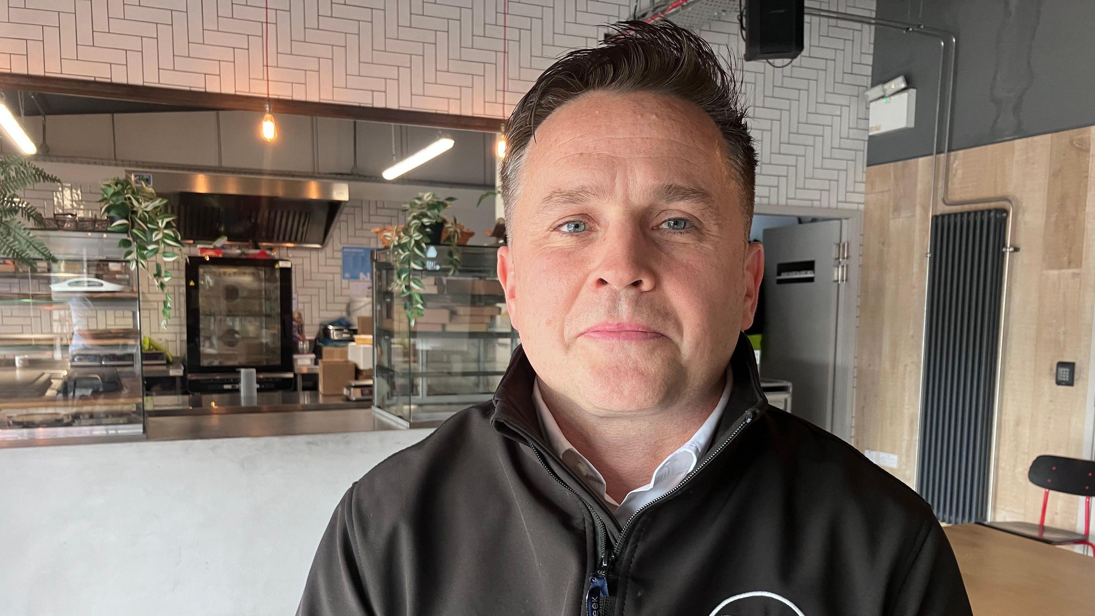 Brian Reid. He has dark hair and blue eyes, wearing a black zip up jacket with white shirt underneath. A cafe counter is behind him.