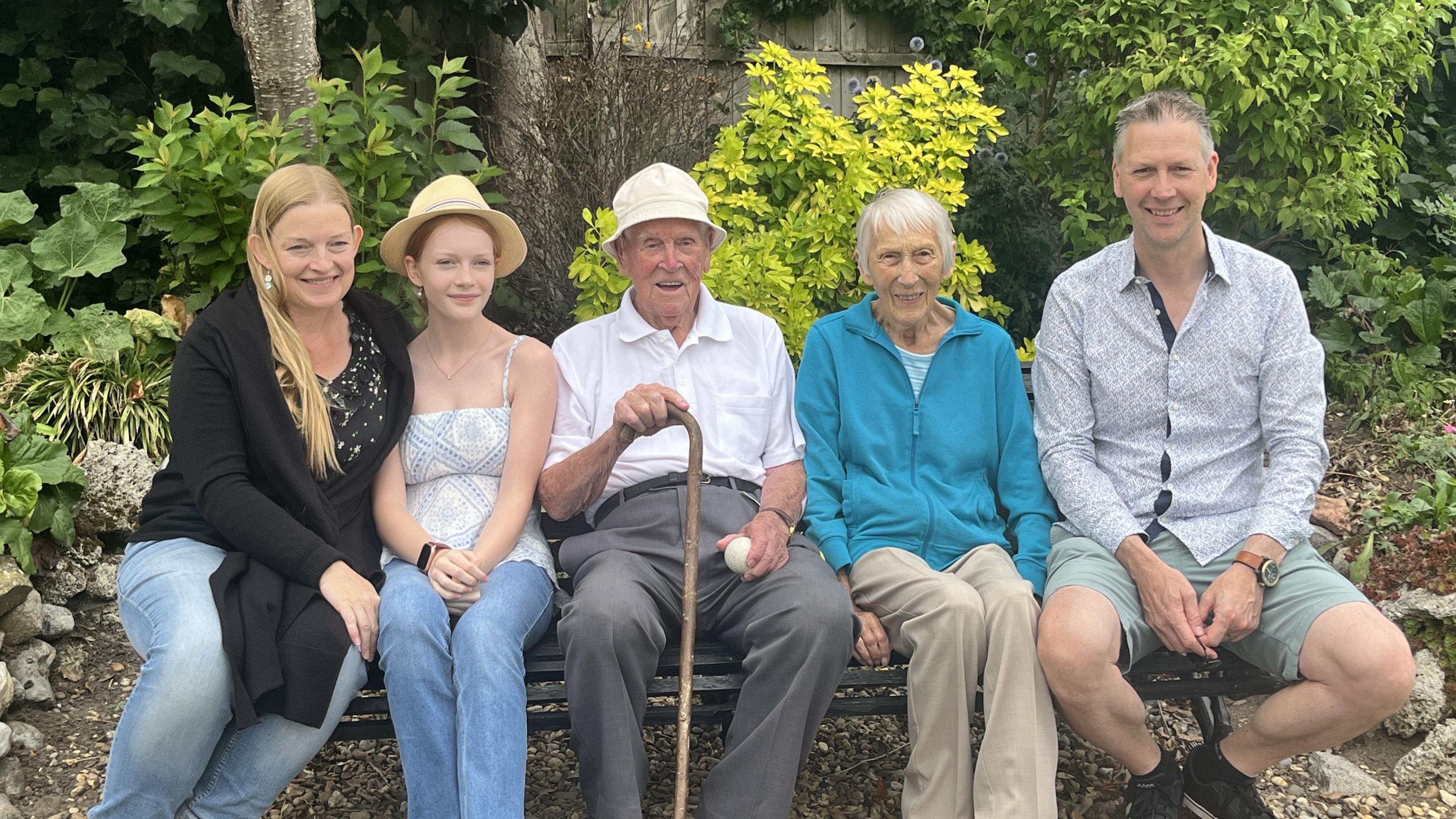 A family of two men, two women and a teenage girl sit on a bench in front of bushes in their garden.