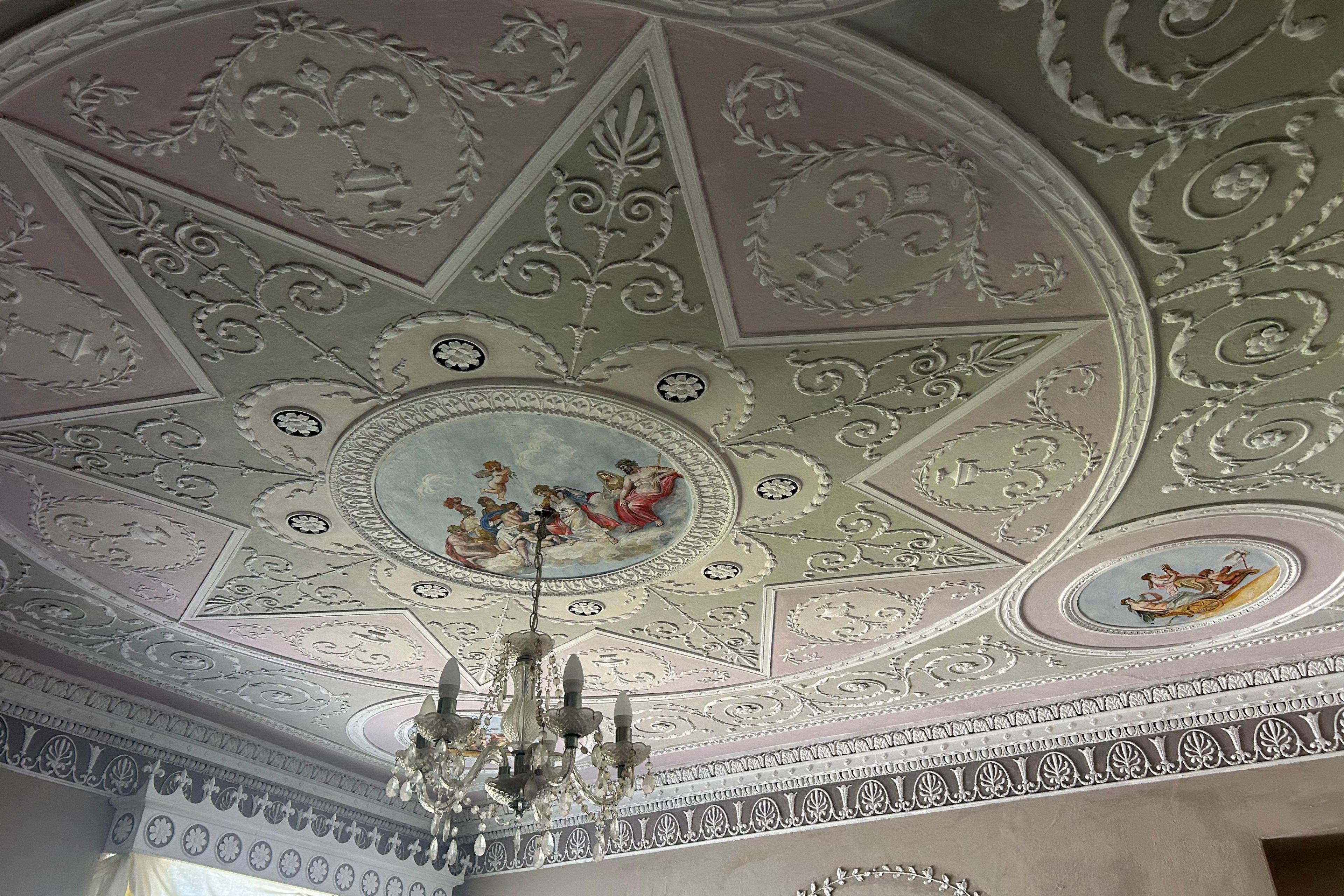 A close up of one of the ceilings inside Mr Jones' home. The ceiling is painted a variety of different pastel colours with intricate white neo-classical detailing. In the centre of the ceiling there is a circle depicting god-like figures sitting on a cloud, with the sky around them. A chandelier hangs from the circle. A large concentric circle is around the artwork, and within that is a smaller star shape, each containing ornate plasterwork lines and patterns. 