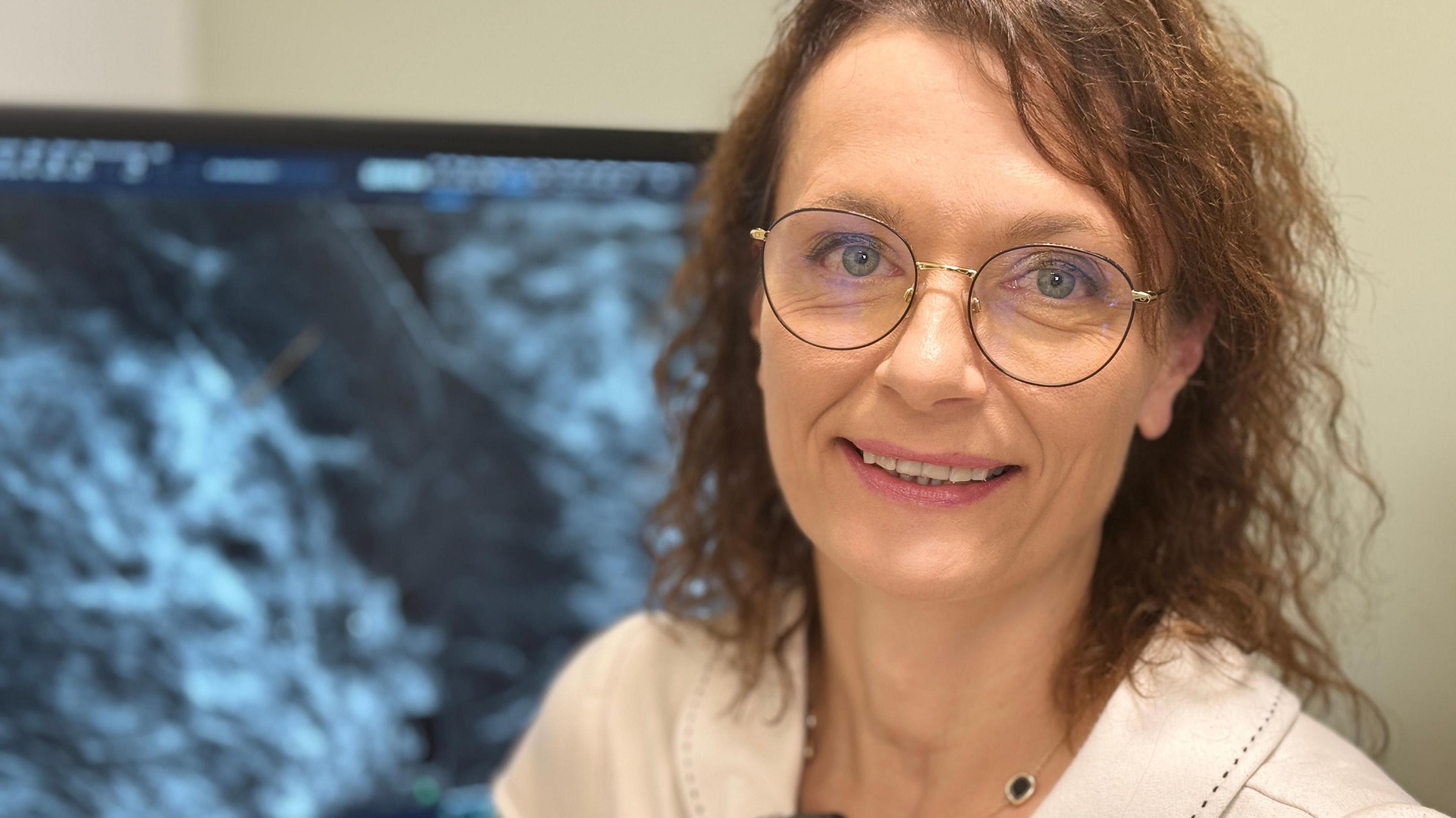 An image of Dr Olga Strukowska looking at the camera. She has shoulder-length wavy brown hair and is smiling. She is wearing a pink top and is in front of a mammogram.