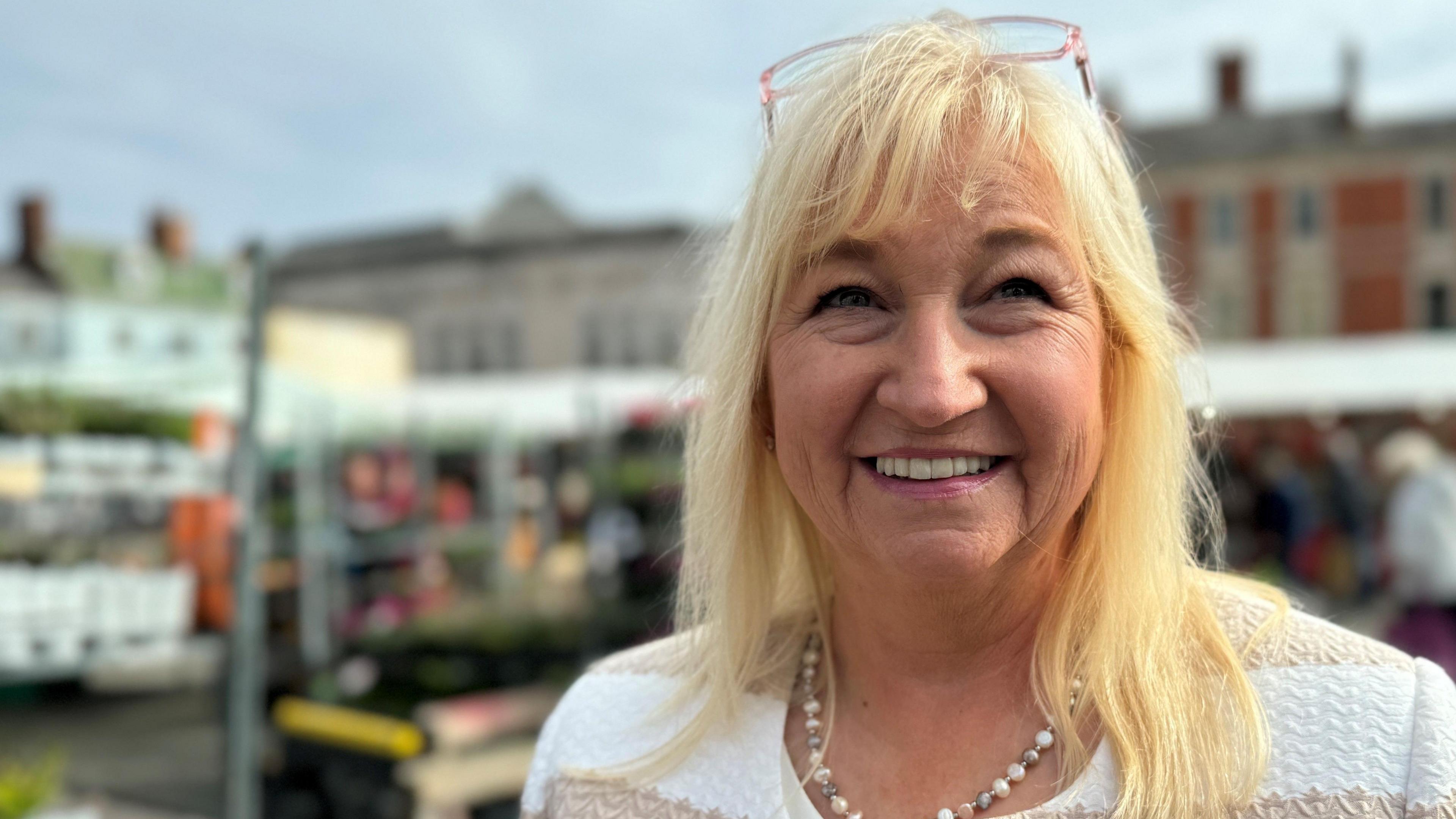 Boston Borough Council leader Anne Dorrian smiles as she stands in front of stalls in the Market Place. She has shoulder-length blond hair, with pink-framed glasses perched on the top of her head. She wears a white and light-brown striped jumper and a matching necklace.