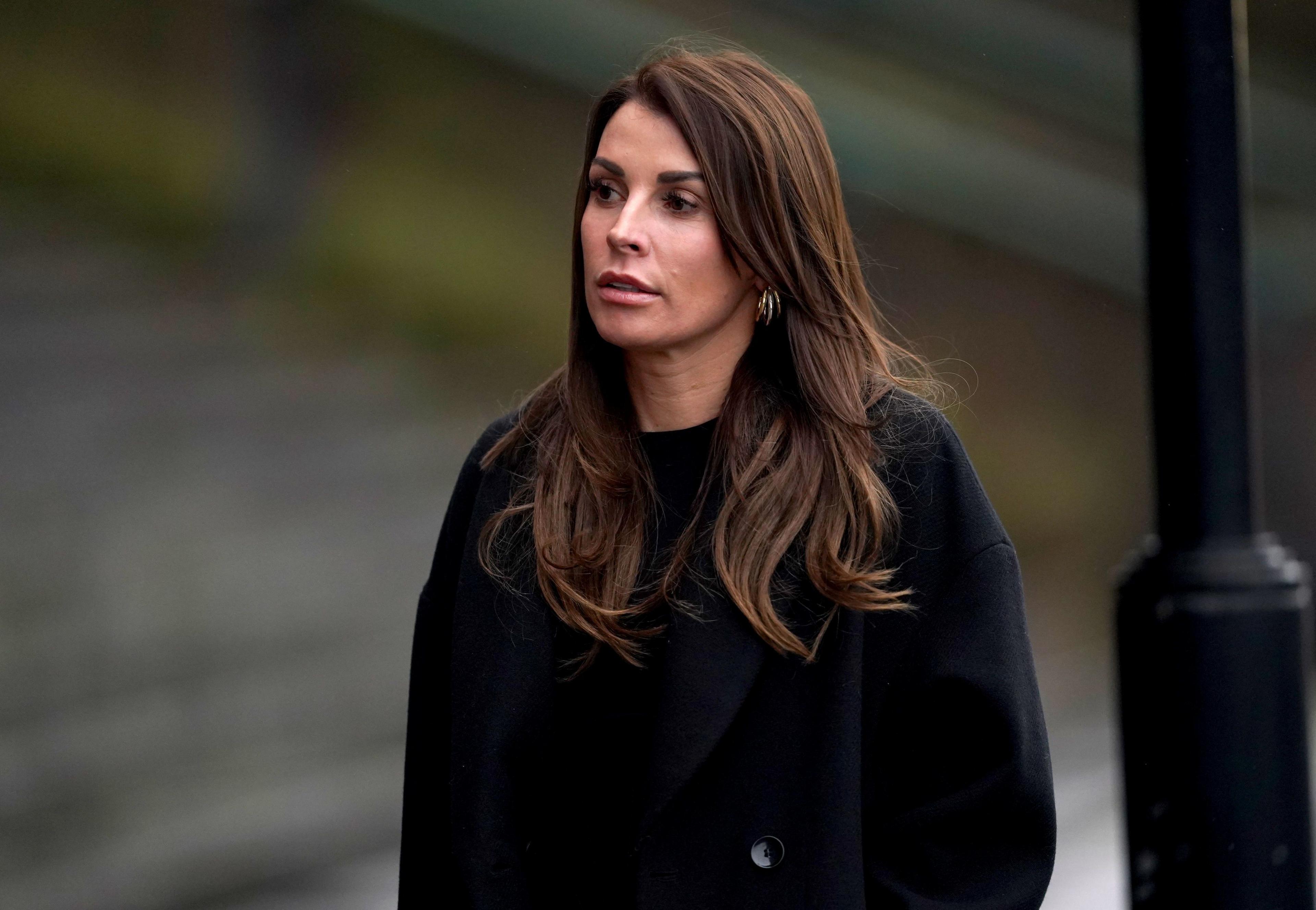 Coleen Rooney is pictured walking alongside a lamppost. She is in a dark jacket and has long, wavy brown hair. 