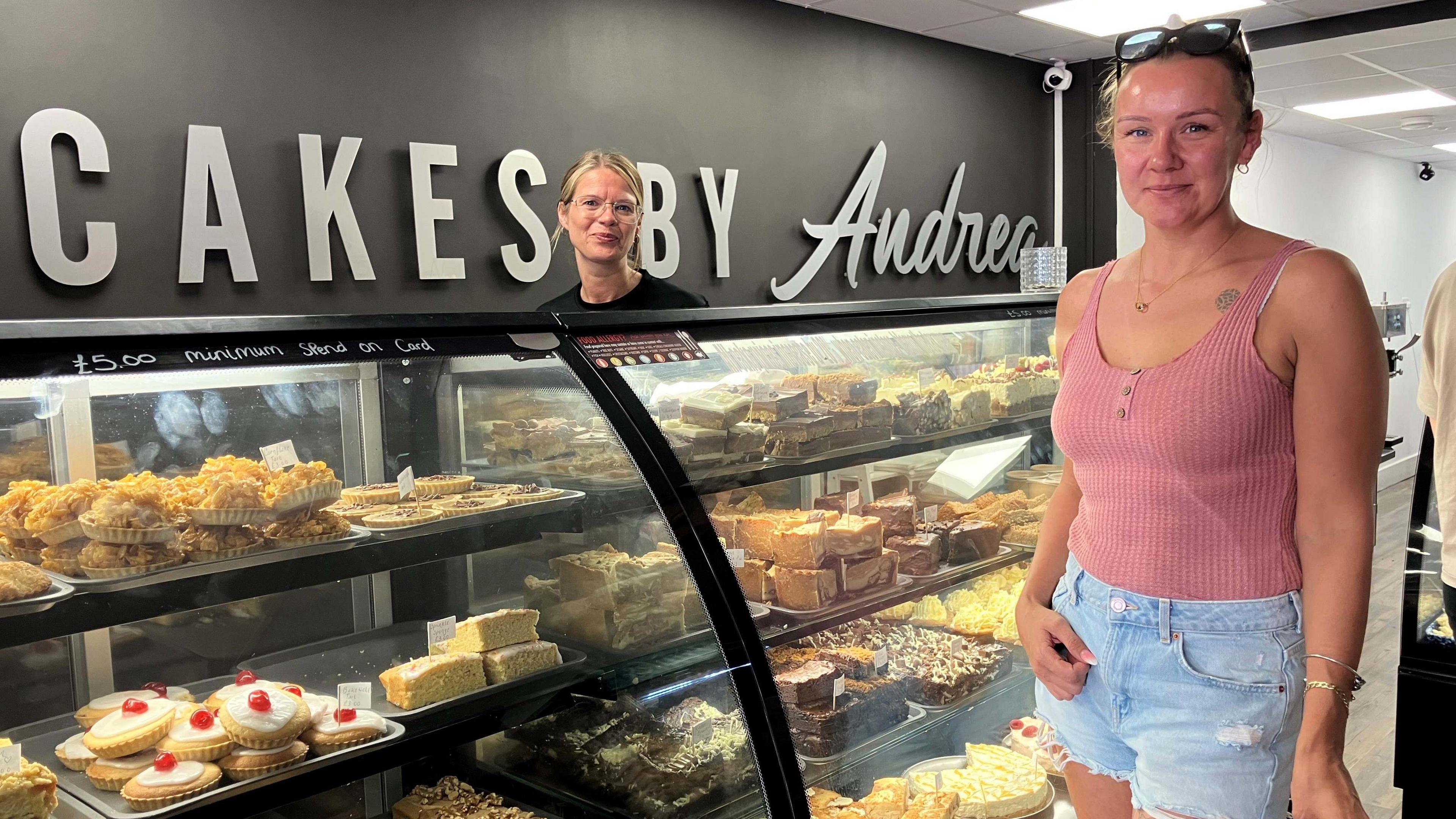 A woman wearing a peach-coloured vest, blue denim shorts and sunglasses on her head is standing in front of a glass cabinet full of cakes. Behind the counter is a woman with long blonde hair and wearing a dark top. A sign says "Cakes By Andrea" on the wall behind them.