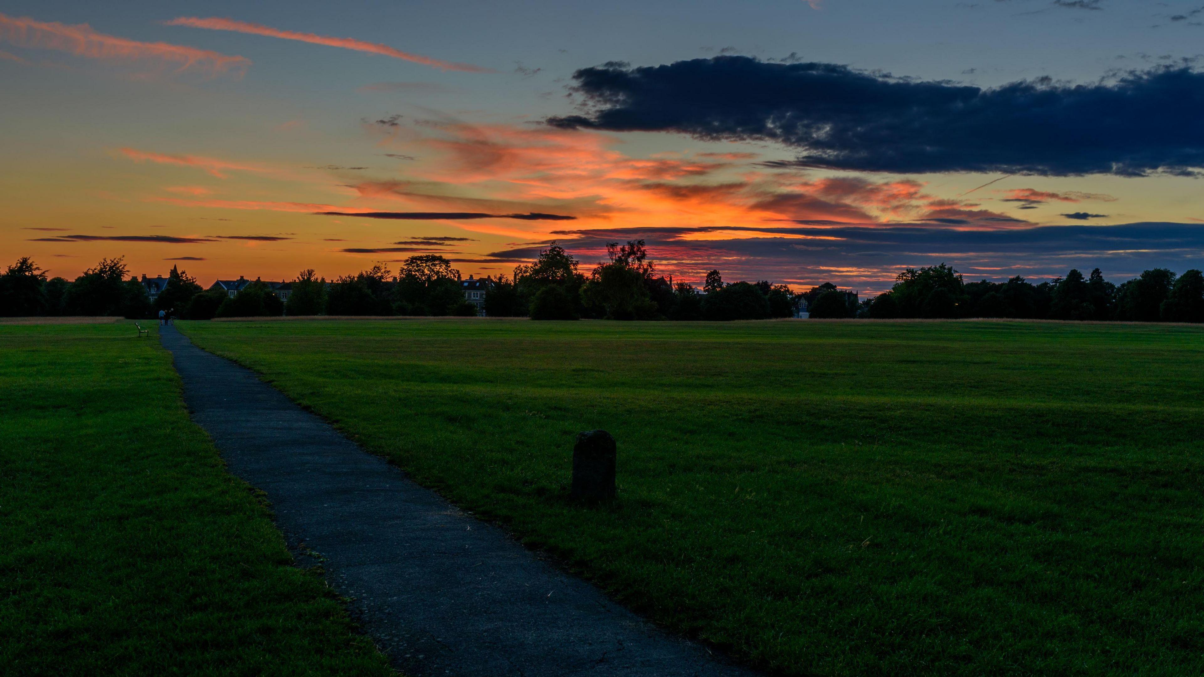The Downs in Bristol at sunset with an orange and dark blue sky. A path can be seen heading away from the camera towards a line of trees and houses
