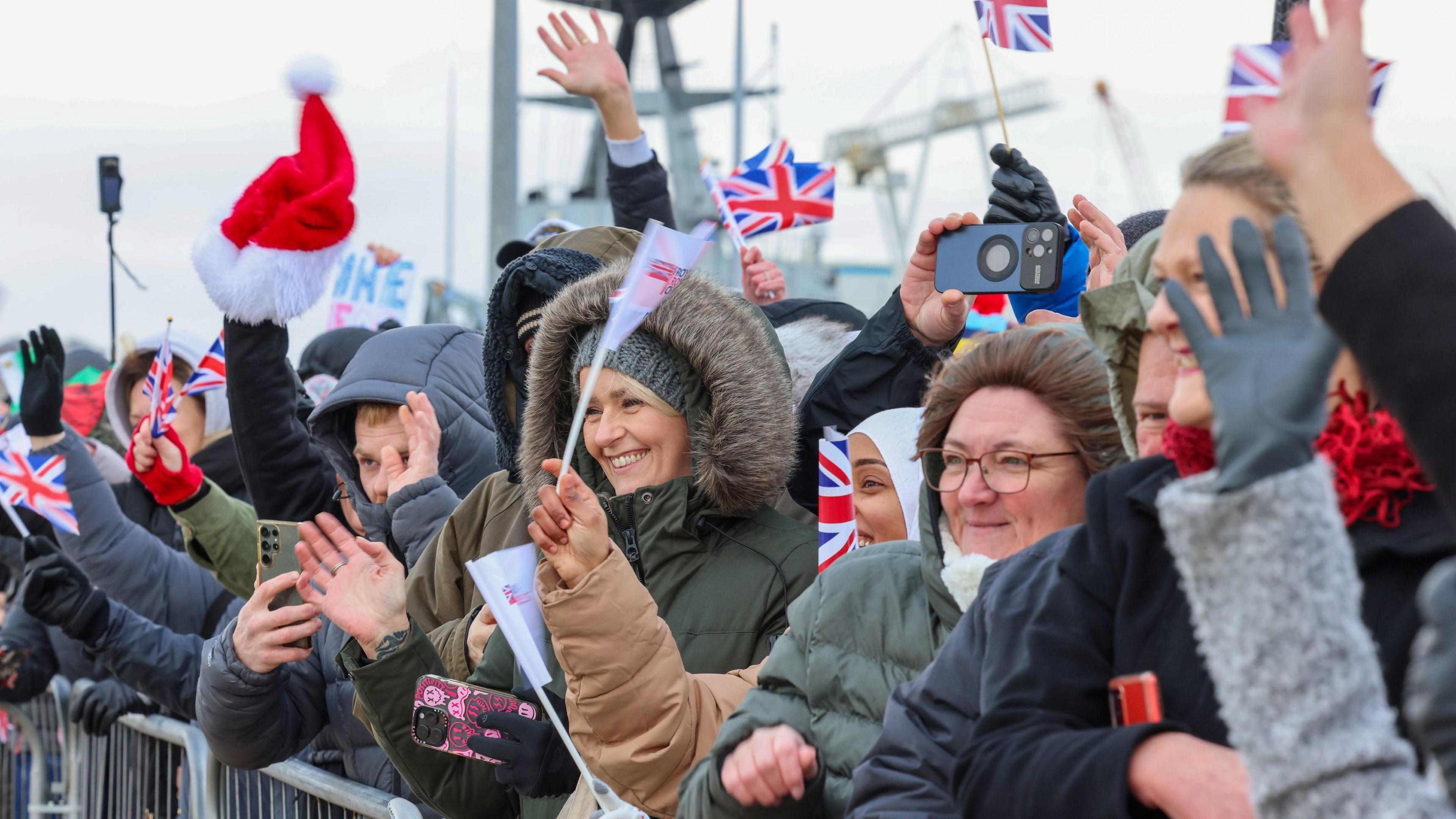 People are lined up against a gate, everyone is smiling and waving as they see HMS Duncan, many are holding Union Jack flags, one person is holding a Santa hat and people are holding up their phones to film the ship entering Portsmouth harbour.