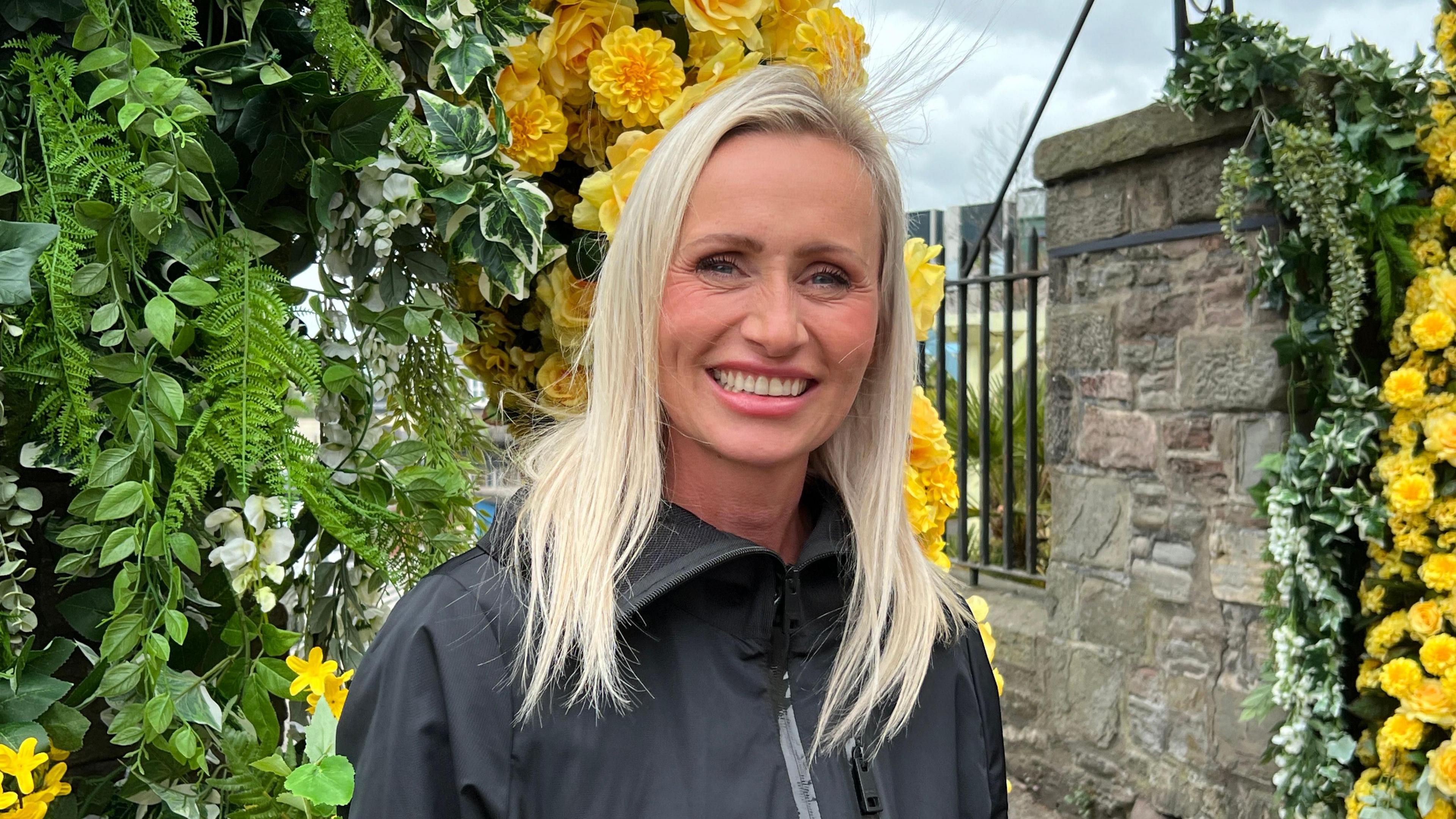 Ms Popplewell wearing a black raincoat standing beside stone pillars where the yellow flower installation is attached. You can see greenery and yellow silk flowers behind her.