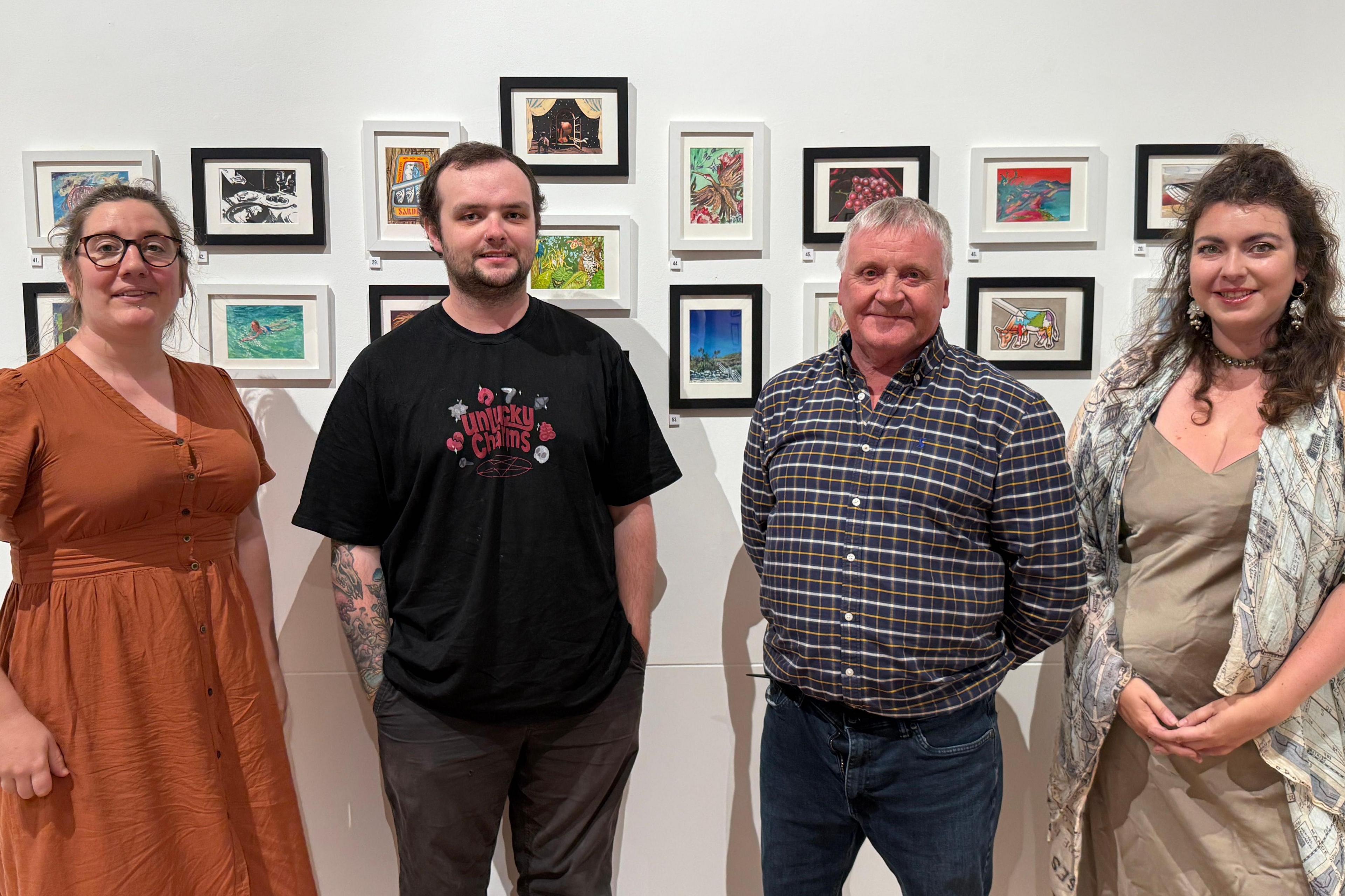 From left to right, a women looking at the camera and smiling. She is wearing glasses and has brown hair which is scraped back. She is wearing a rusty brown colour dress. A man looking at the camera and smiling.He has short dark hair and a short beard. He is wearing a black t shirt with red writing on. He has his hands in his pockets. A man looking at the camera and smiling. He has his hands behind his back. He has grey hair and is wearing a long sleeved patterned shirt and blue jeans. A women looking at the camera and smiling. She has her hands clasped together by her stomach. She is wearing a silk style long dress and a shoal that has a vintage map of Paris print. She has medium length brown hair.