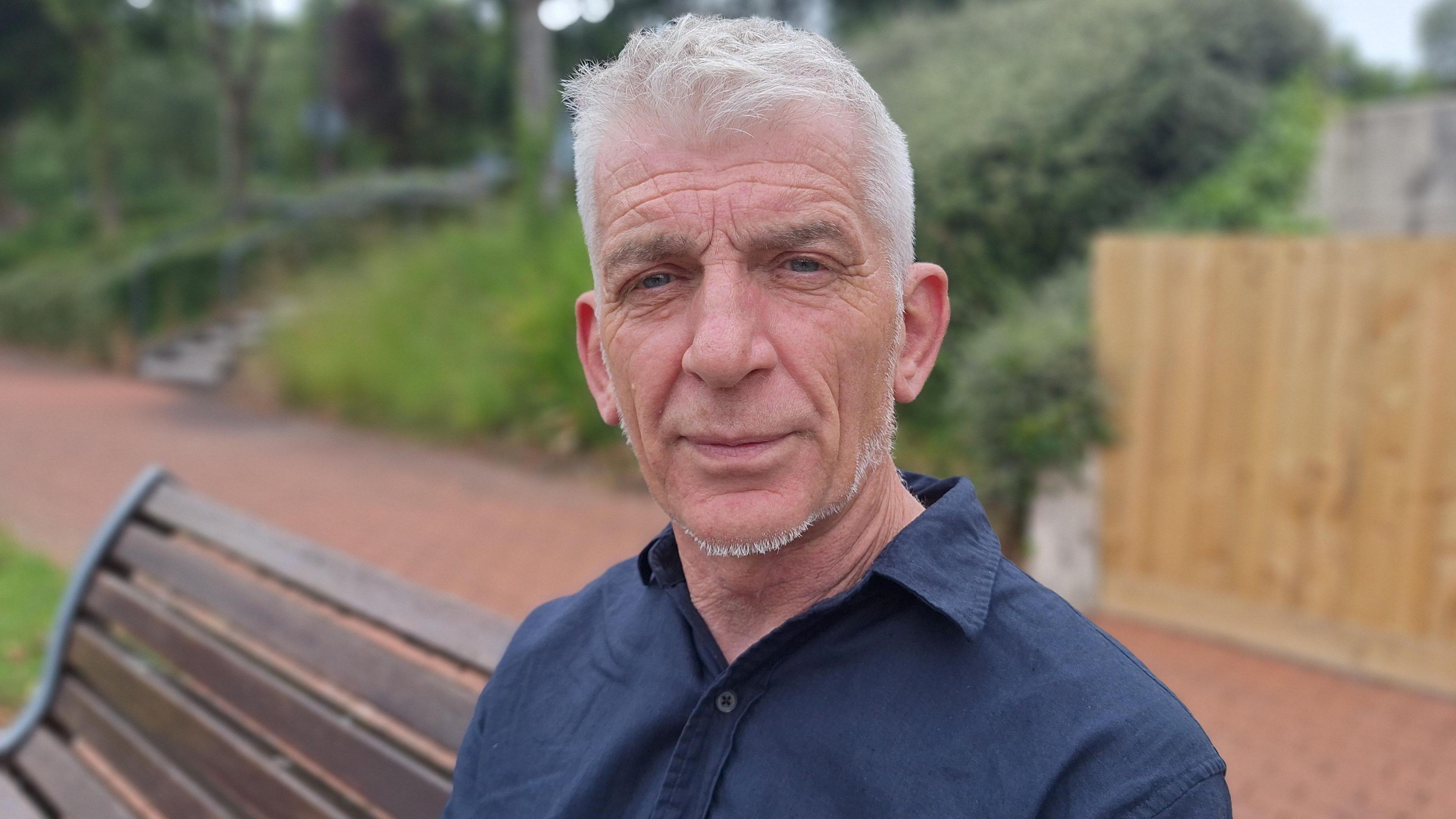 Stuart Todd has short white hair and a slight beard. He is sitting on a bench on a university campus, looking into the camera. He is wearing a dark blue shirt and there are trees and plants behind him. 