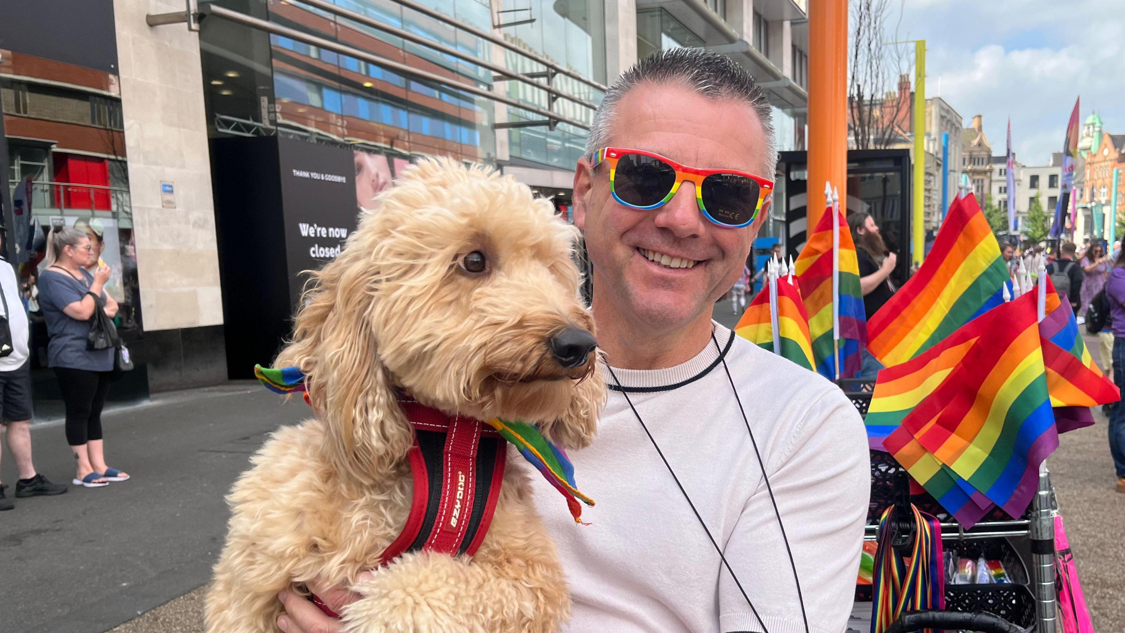 A man wearing rainbow-framed sunglasses and holding a dog which is wearing a rainbow tie on its harness. 