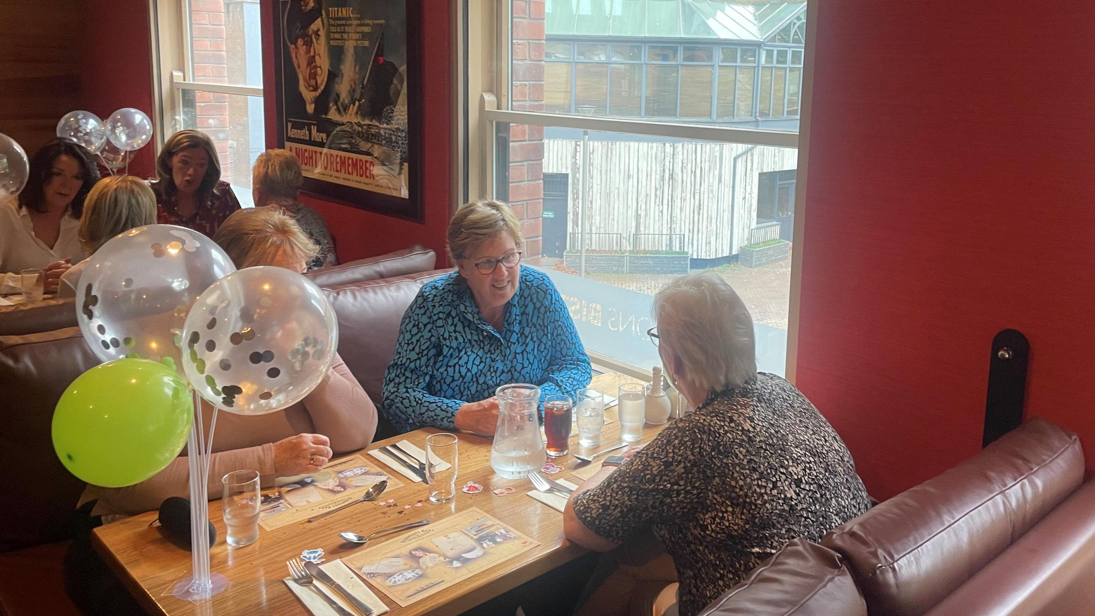 Three women sit at at a table one is blocked from the camera by translucent balloons, two sit by the window facing each other. One is in a blue and black top, the other (with her back to the camera) is wearing an brown-coloured top