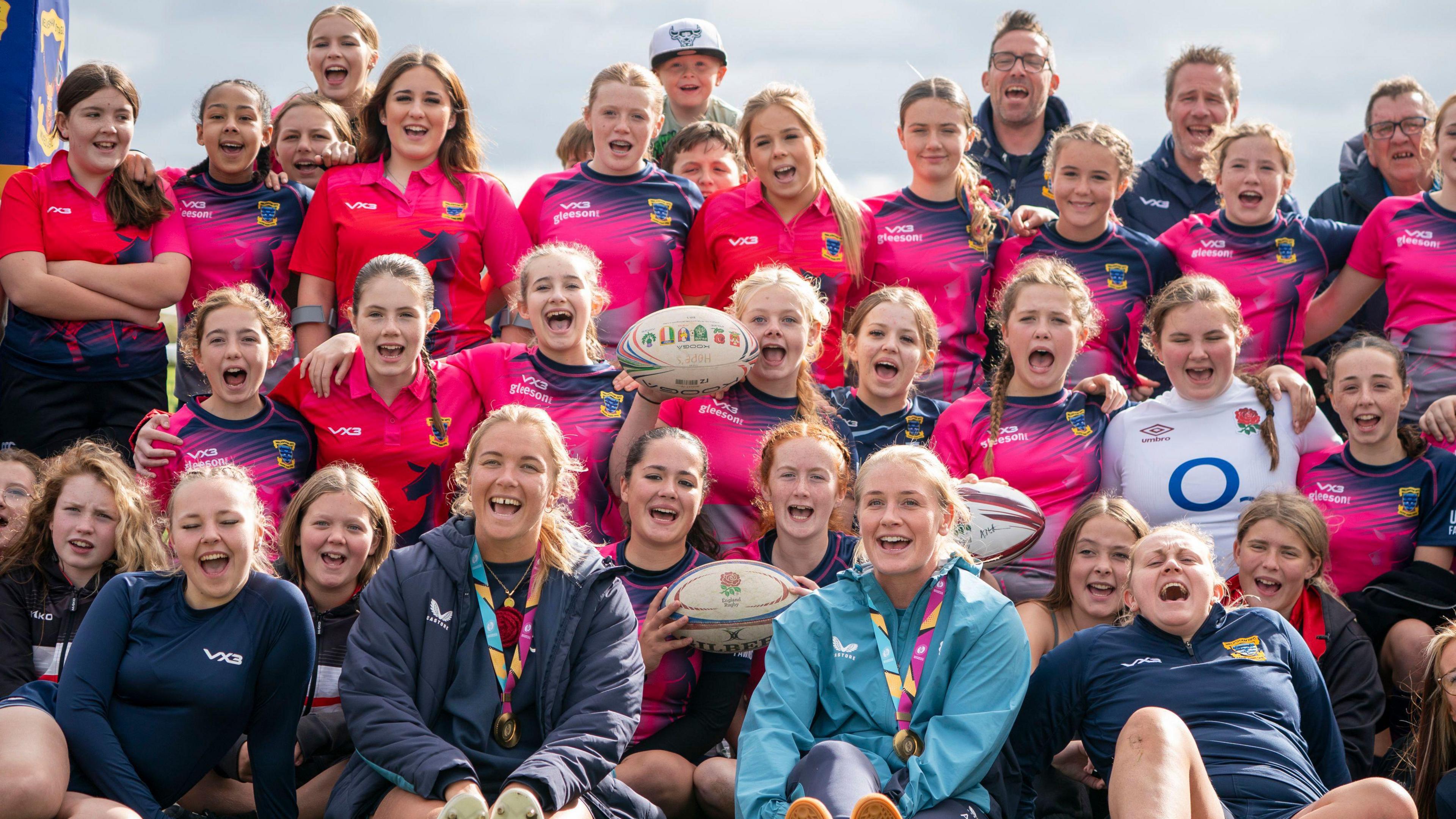 Dozens of young female rugby players group together and cheer as they look at the camera. Lined up sitting down in front of them are England players Zoe Aldcroft and Alex Matthews