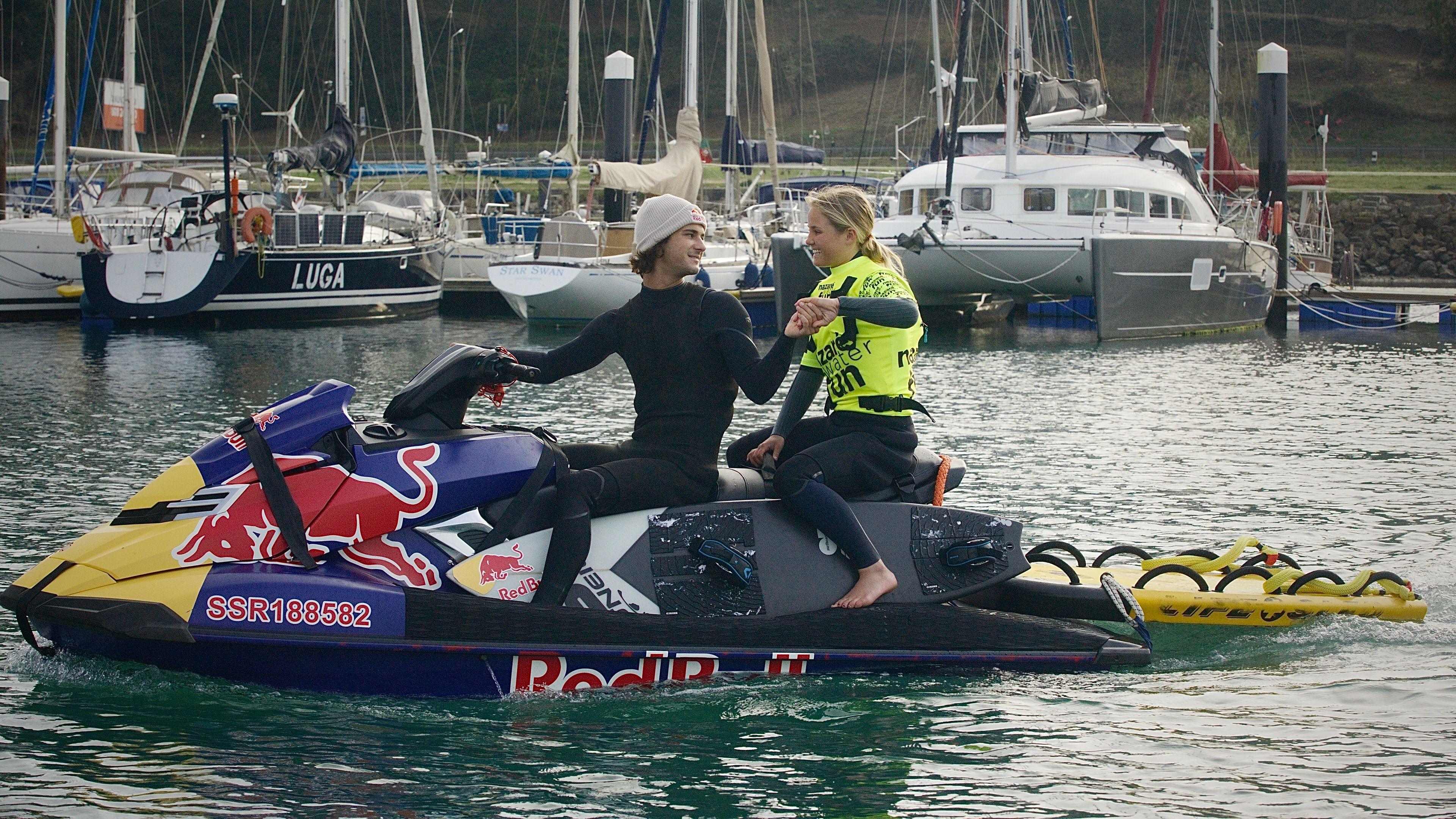 A man and a woman on a Red Bull branded jetski. The sea is green and they are in a harbour. The jetski is towing a yellow floating object which is a safety sled. The man is wearing a black wetsuit and is shaking the hand of the woman sitting behind him. She is wearing a green top over a black wetsuit. 