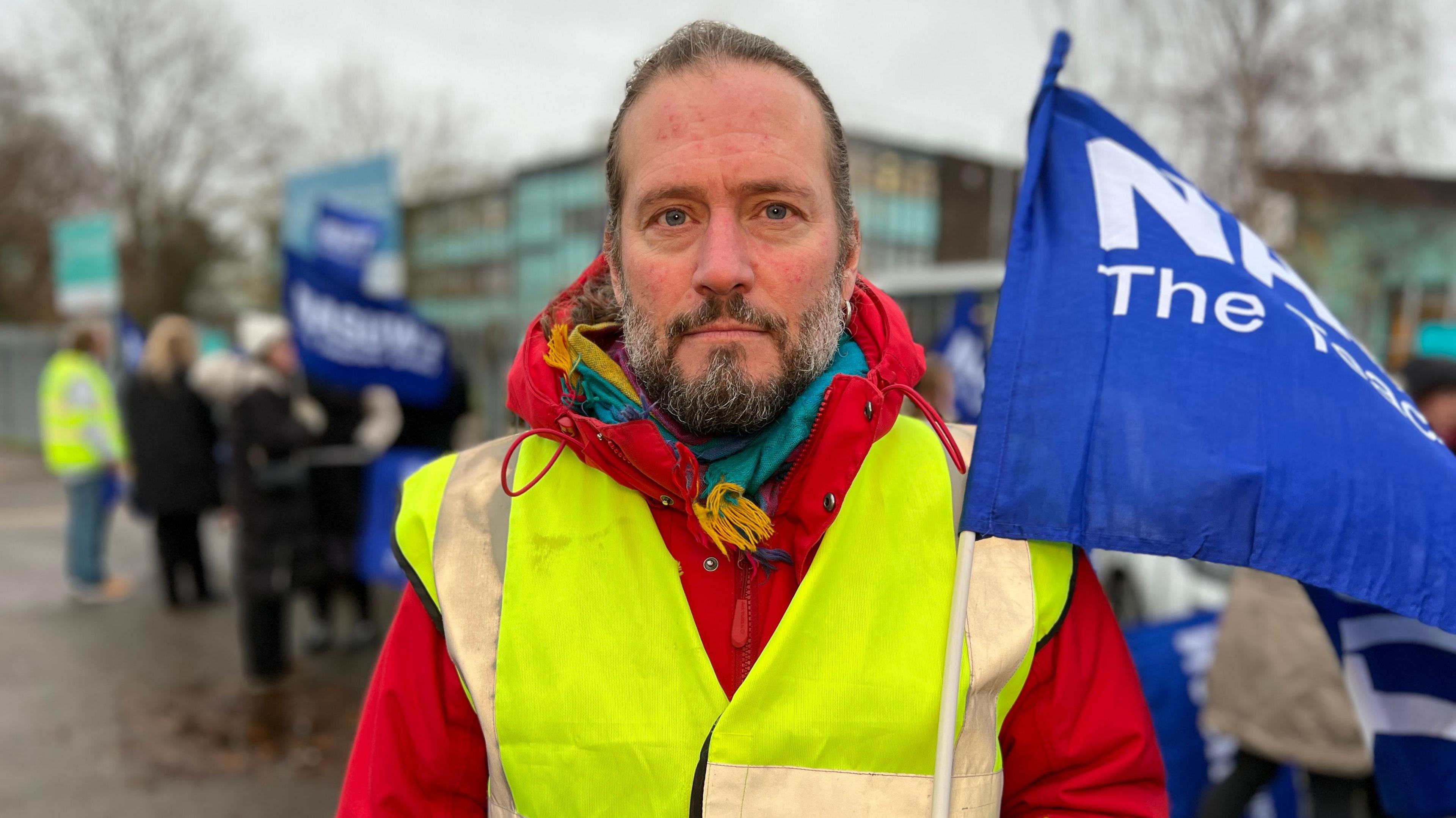 Kim Vollerthun in a high visibility jacket, with a red coat underneath. He has a beard and his hair appears to be tied back in a pony tail. He is holding a blue flag. There are people crowded behind him.