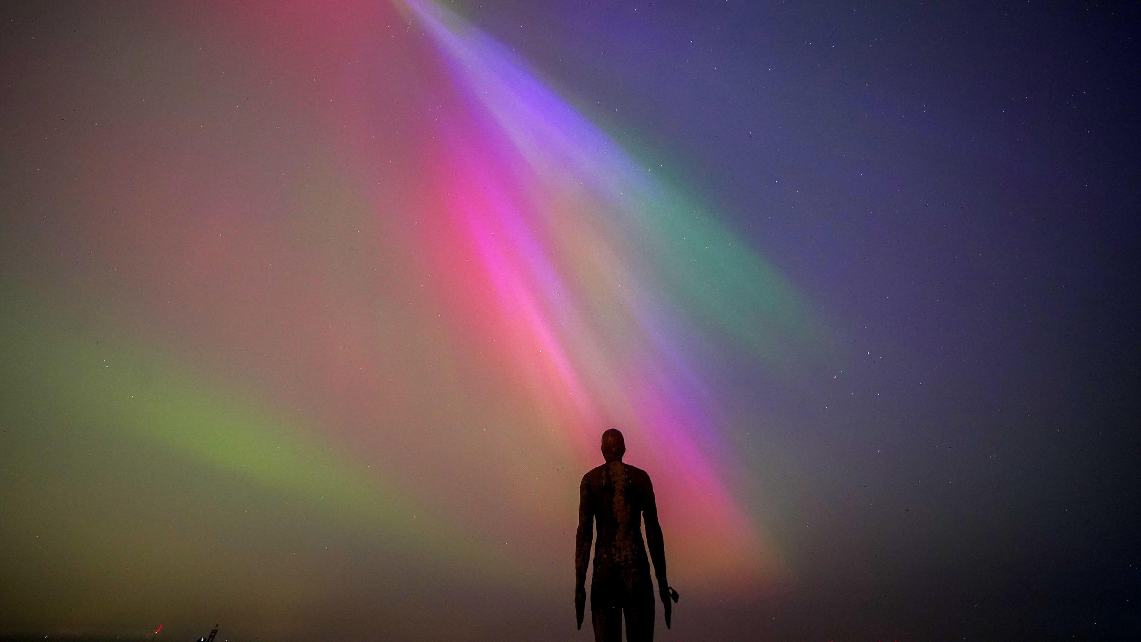 An iron statue with its arms by its sides is pictured in silhouette underneath the glorious pinks, greens and blues of the Northern Lights.