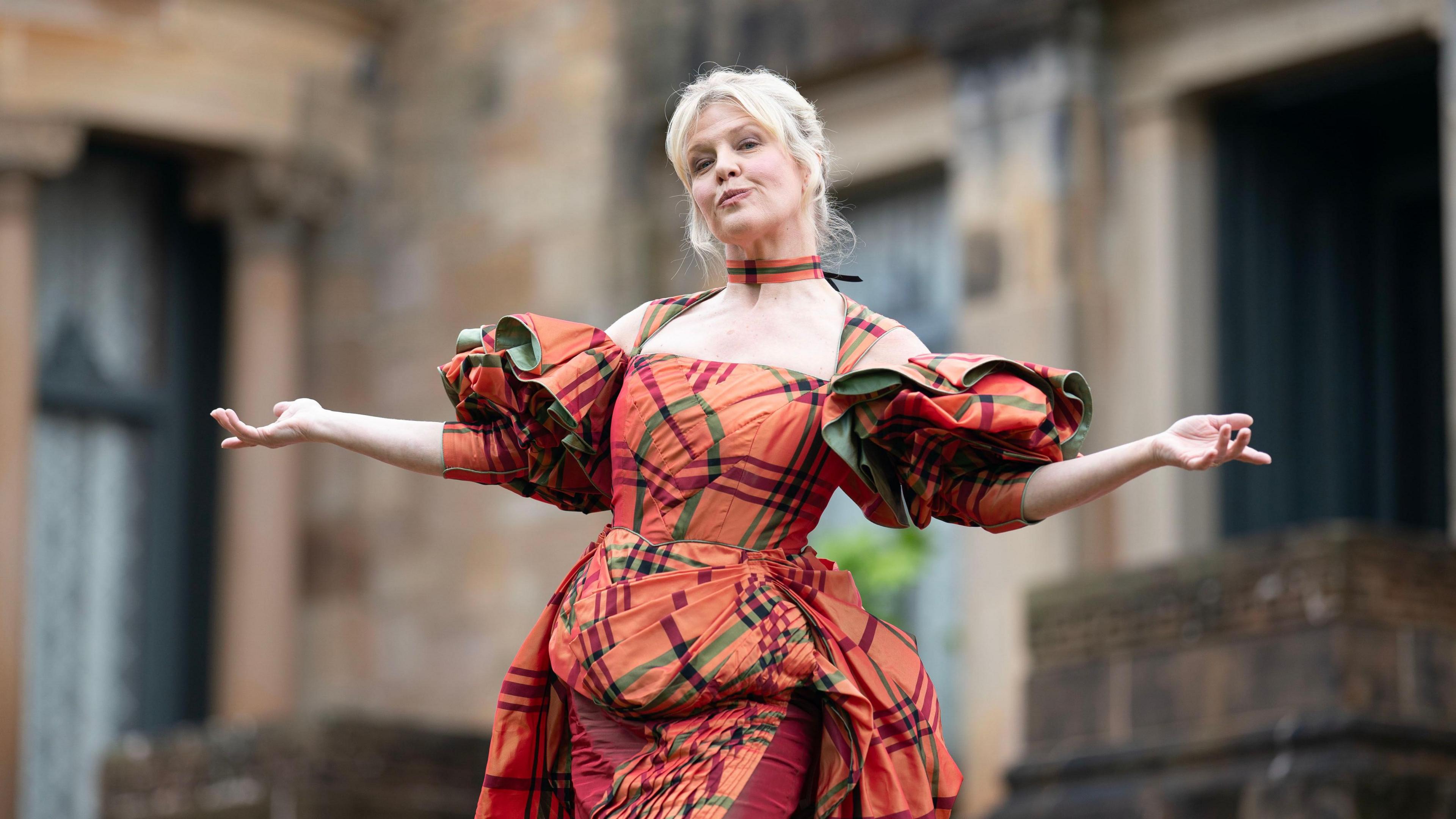 A woman, Ashley Jensen, with blonde hair poses in front of a grand looking building. She has both her arms outstretched and is pouting. She is wearing a costume dress which is orange and green tartan.