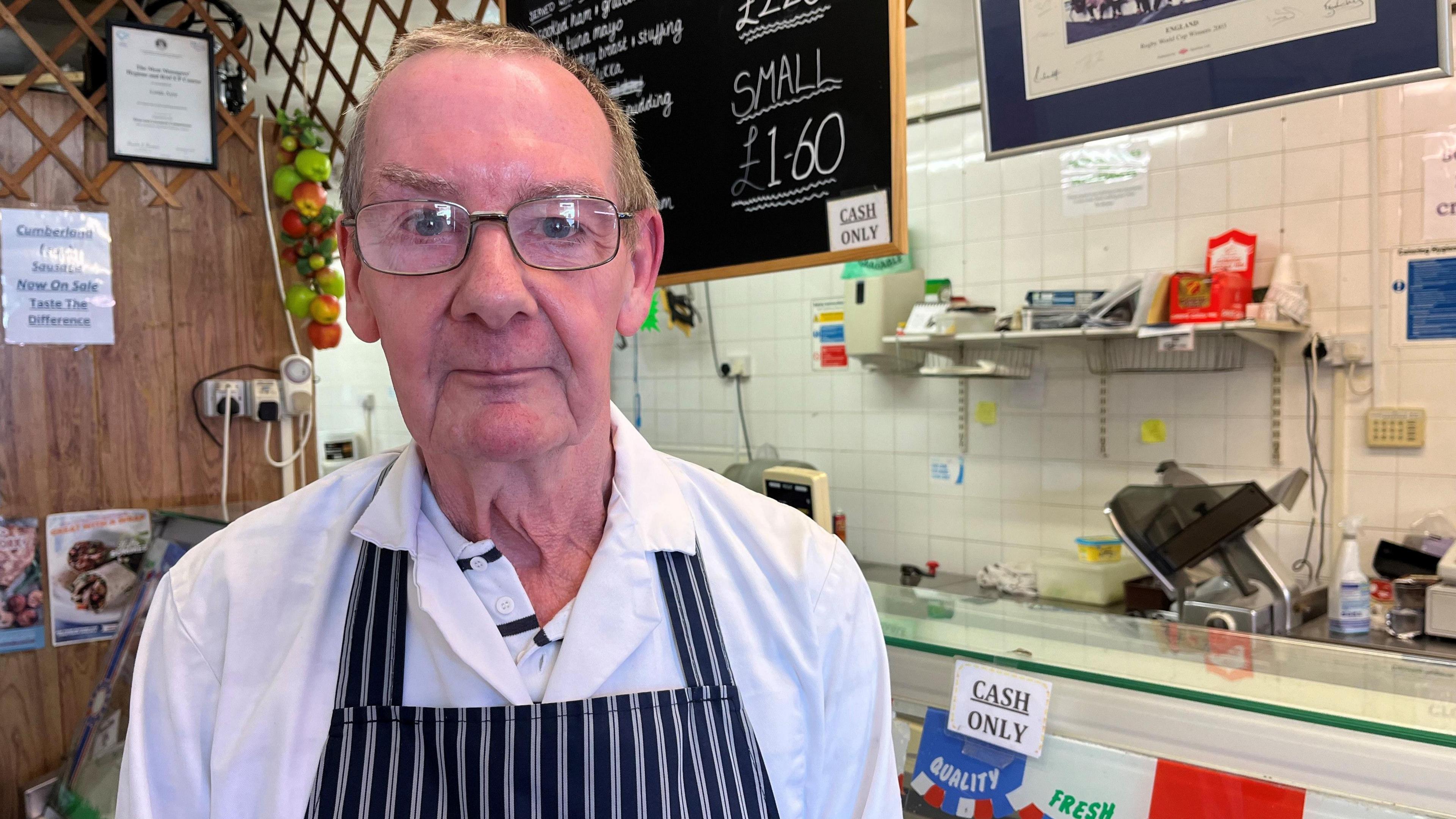 A photo of Michael Ayre, a man with short grey hair and glasses wearing a stripped apron and white shirt. He's standing in front of a counter which says 'cash only'