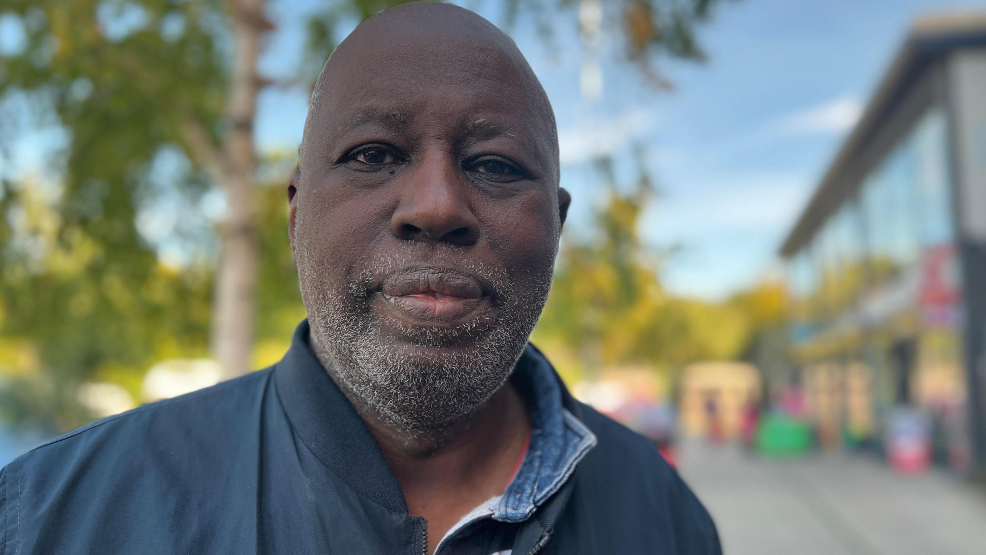 A man looks directly at the camera as he is photographed on a street with shops in the background, dressed in a blue denim shirt and navy jacket. 