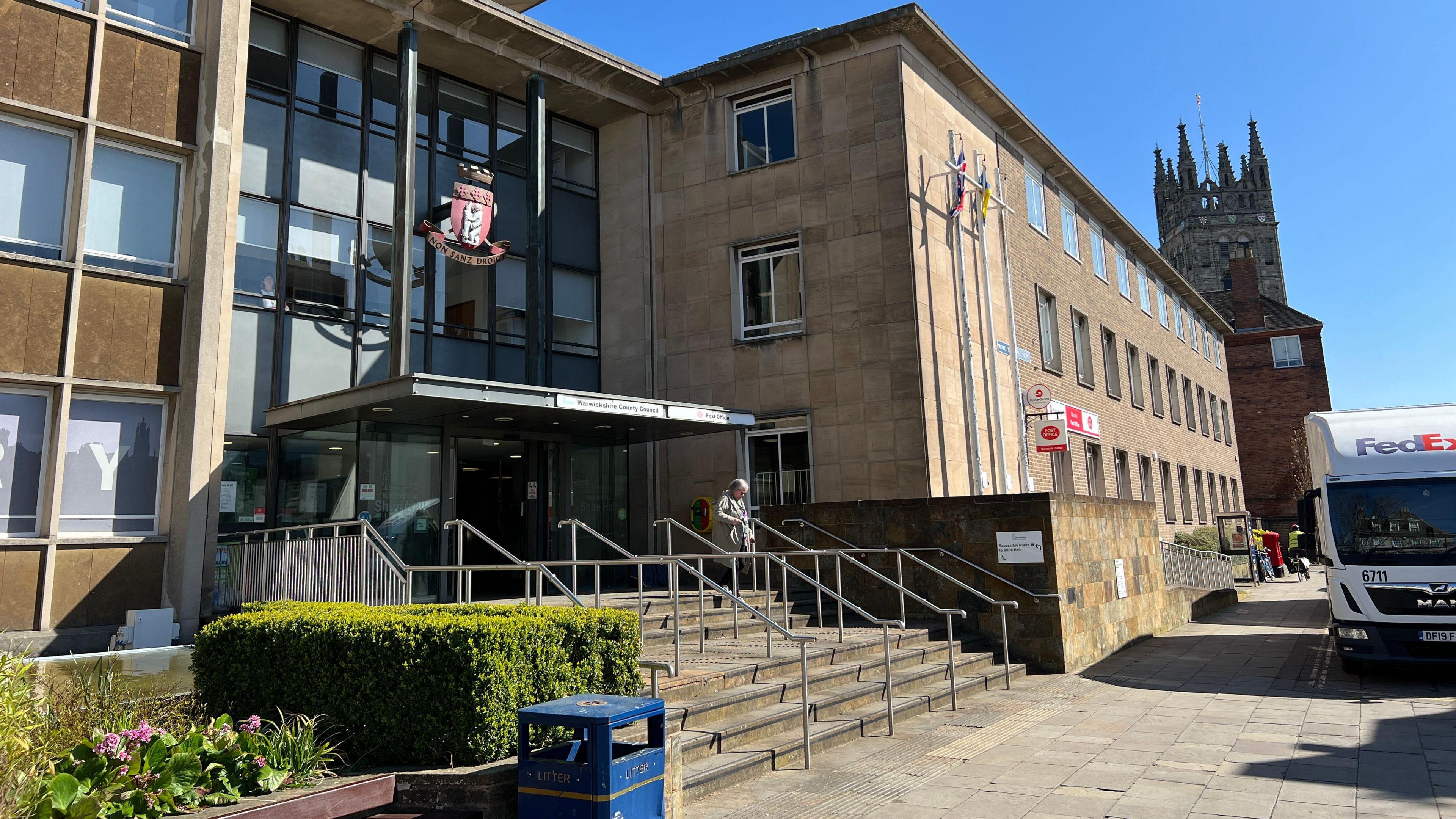 The outside of Shire hall, a stone and glass building with a canopy over the front door topped with a red and white shield.