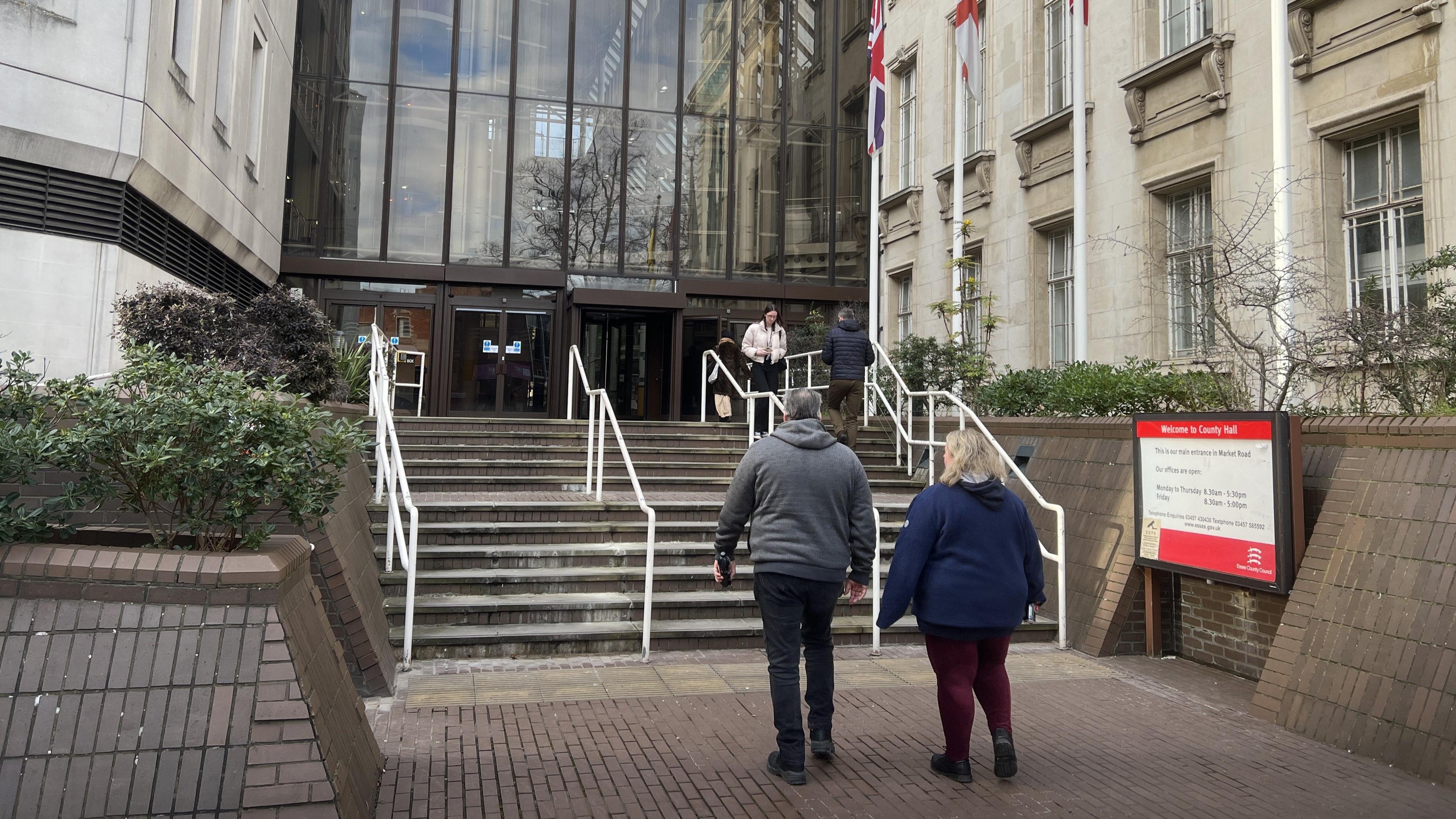 A man in a hoodie and and woman in dark red leggings make their way to a large grey stone building's steps.