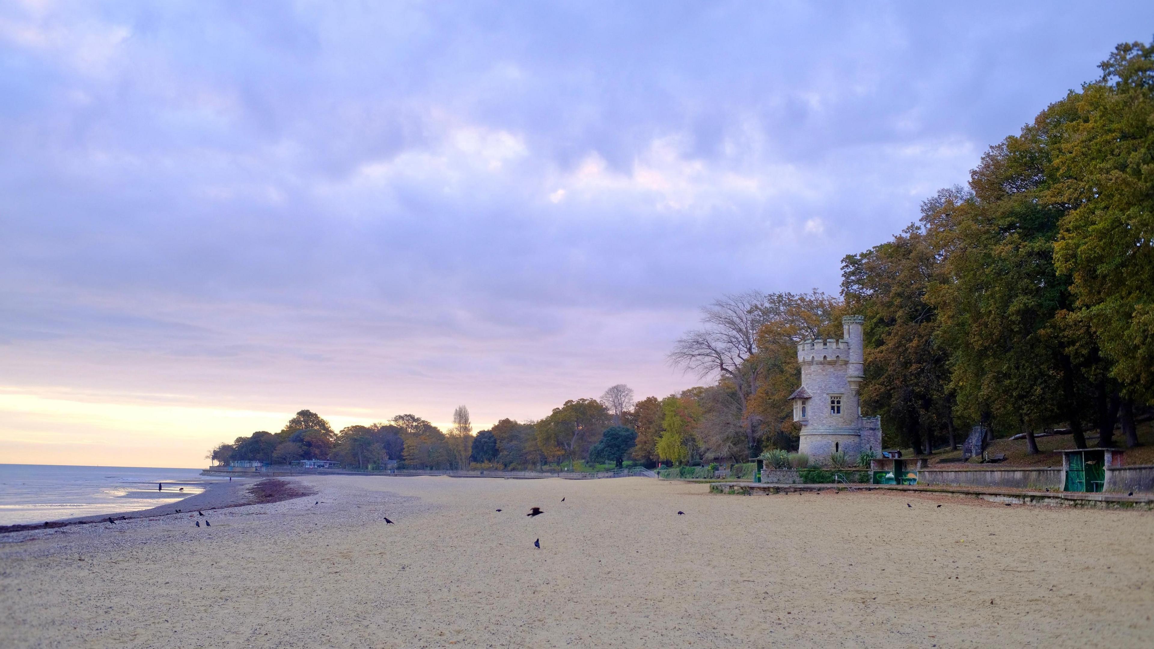 Ryde beach on the Isle of Wight. The wide sandy beach lined with trees with autumnal colours. There is a small castle-style stone tower on the shoreline.